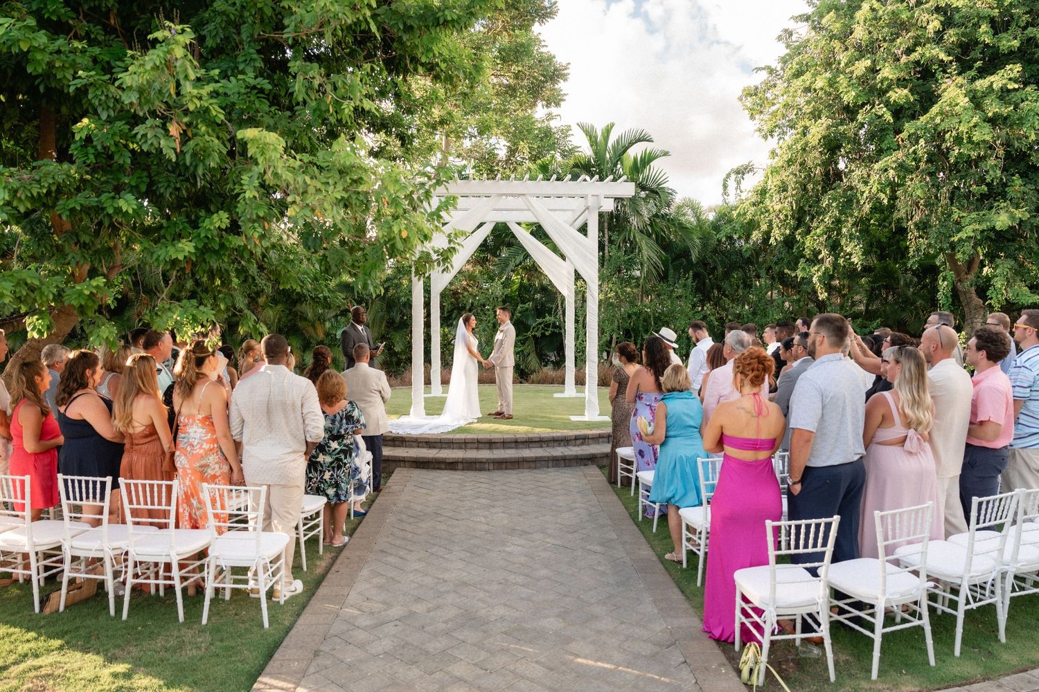 Wedding ceremony setup with white chairs on a garden path leading to a white arch altar surrounded by lush tropical foliage.
