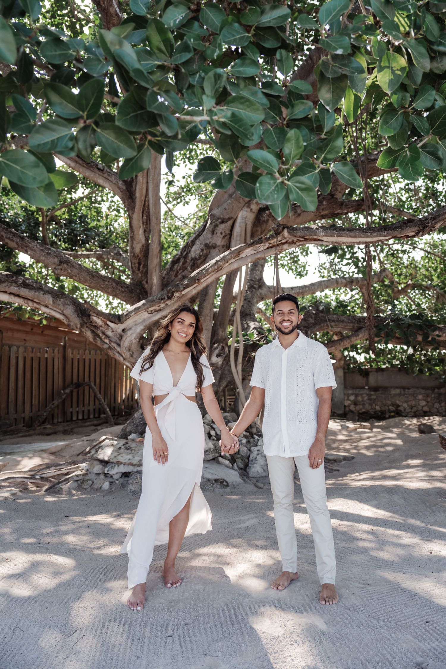 A couple in matching white outfits poses under a large tropical tree with lush green foliage.