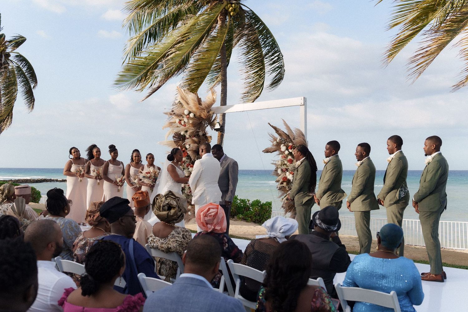 Wedding ceremony setup on a beach with palm trees and guests seated on white chairs facing an arch decorated with flowers.