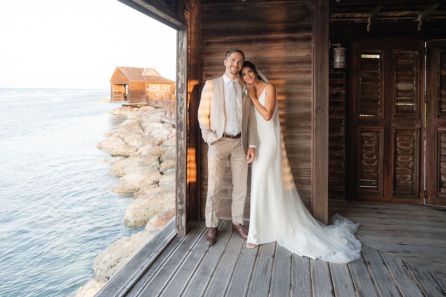 Newlyweds pose in rustic wooden structure overlooking ocean at sunset.