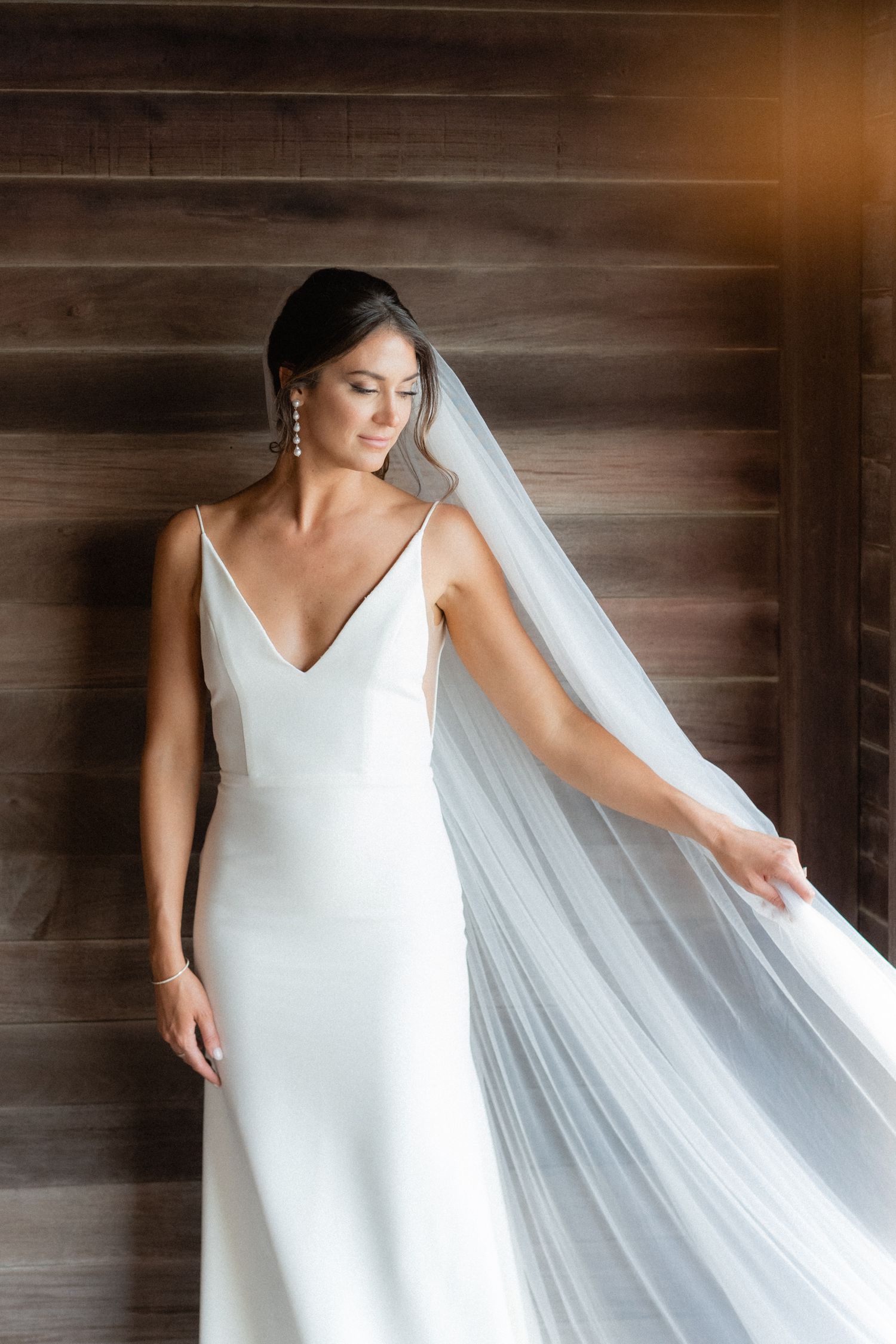Elegant bride in minimalist white gown with flowing veil poses against wooden wall.