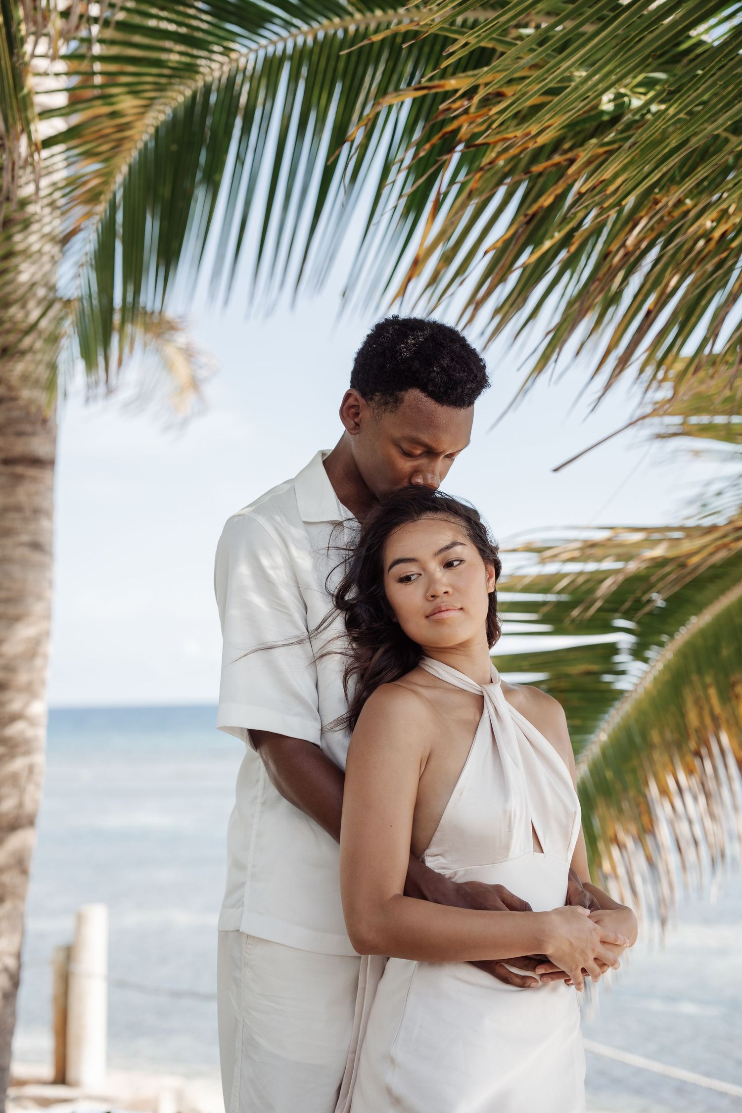 A romantic couple in white clothing embrace beneath palm fronds on a tropical beach.