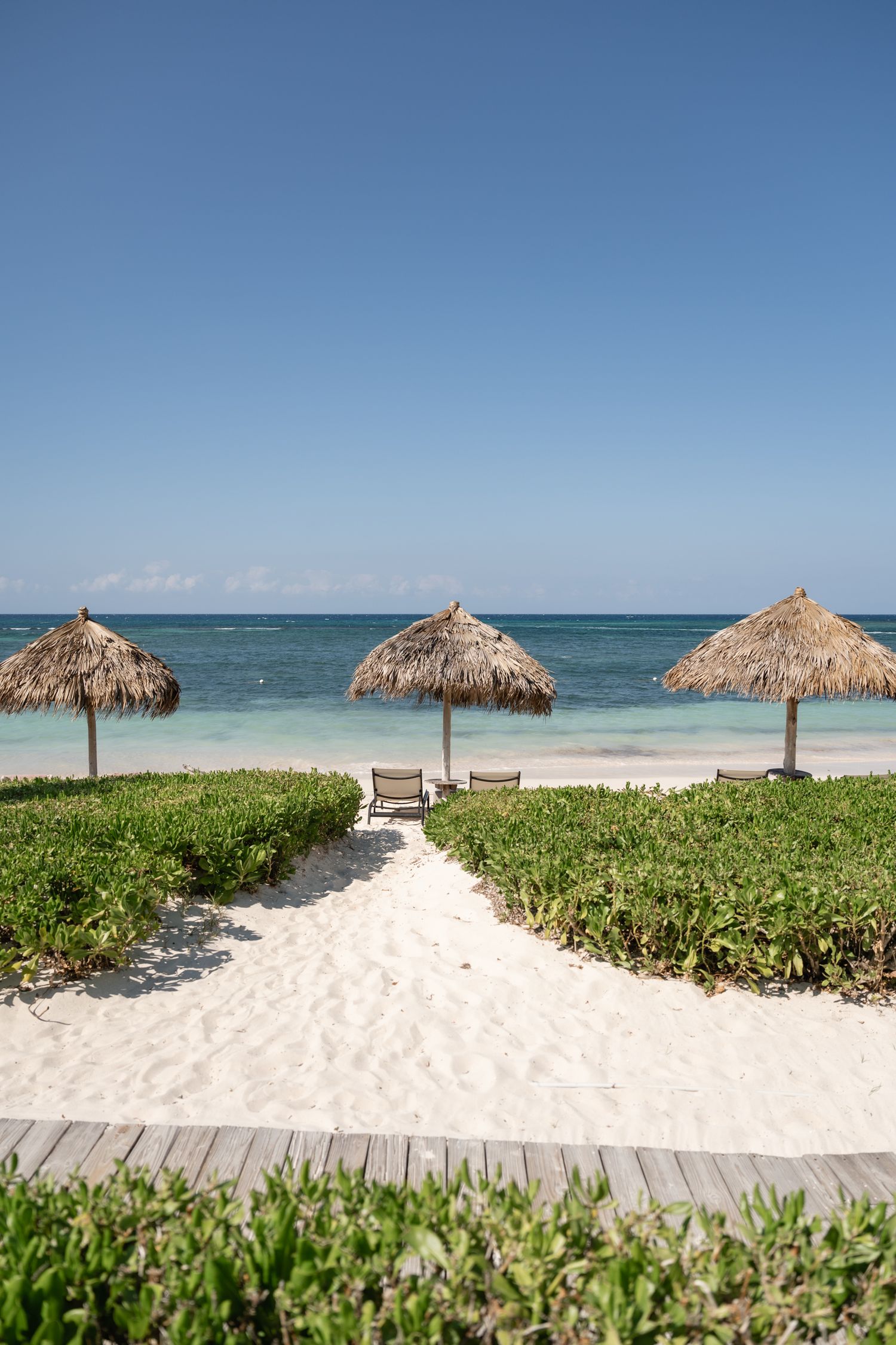 Three thatched umbrellas line a pristine white sand beach with wooden walkway and tropical vegetation.