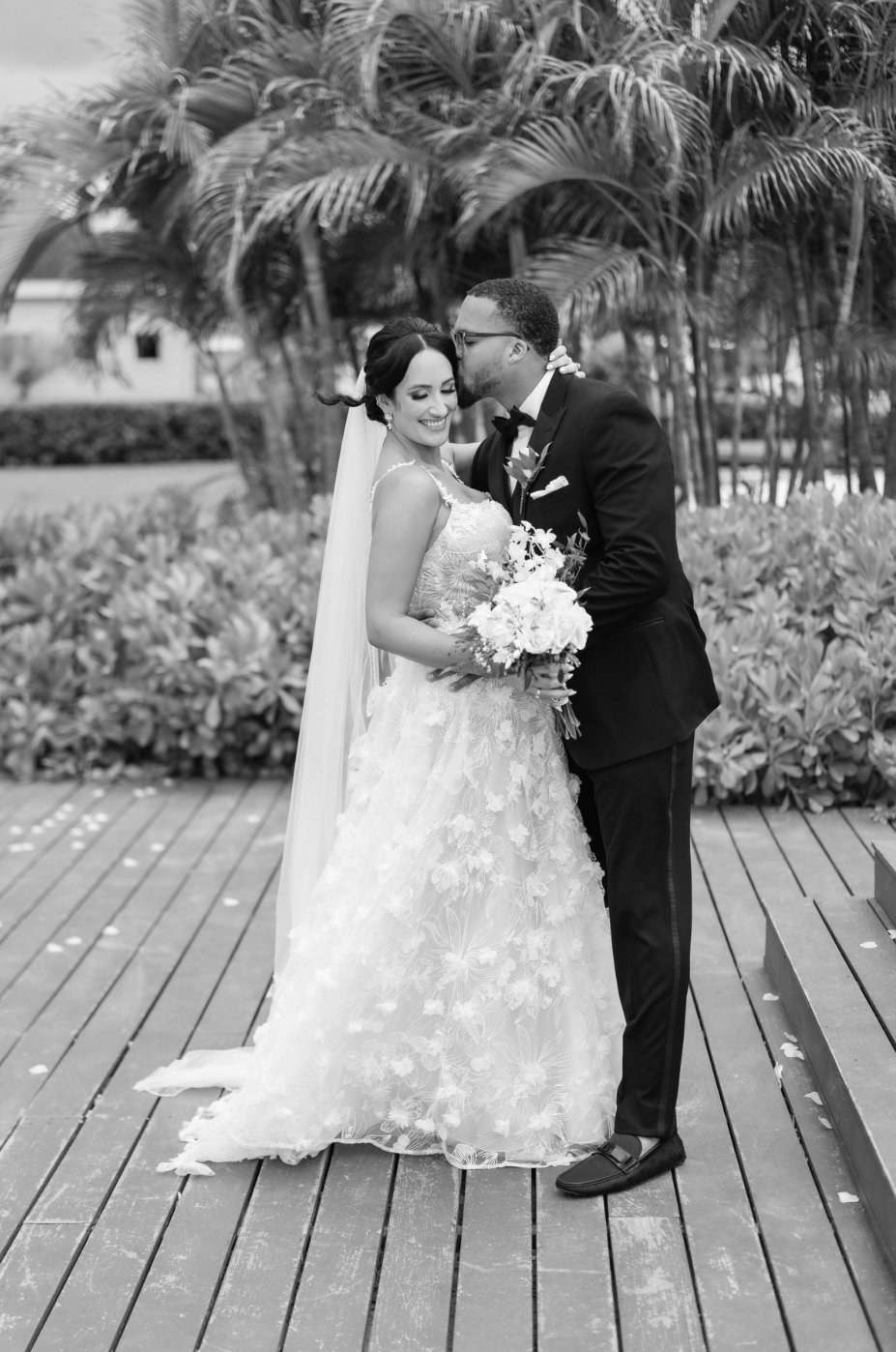 A romantic wedding couple shares an intimate moment on a wooden deck surrounded by tropical palm trees.