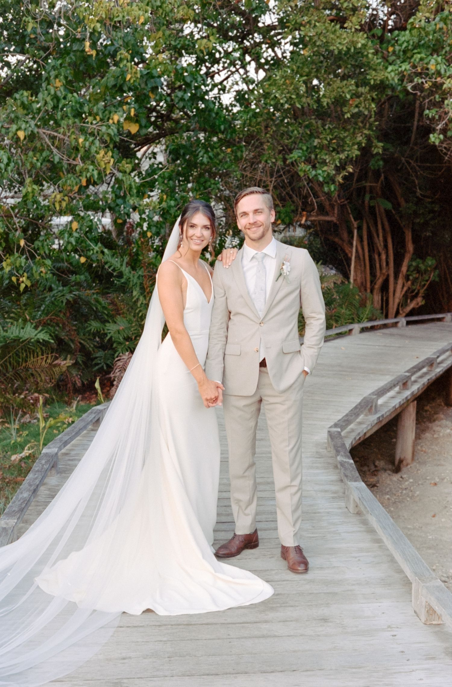 A newlywed couple poses on a wooden boardwalk surrounded by lush greenery.