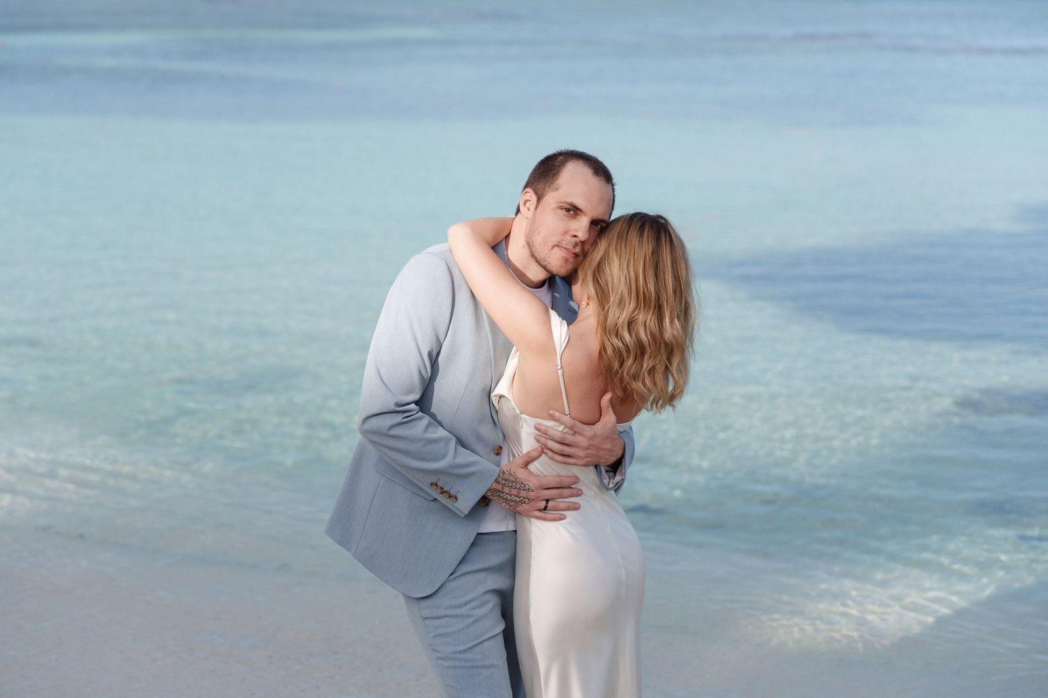 Couple in formal wear sharing intimate moment on beach with turquoise water background.