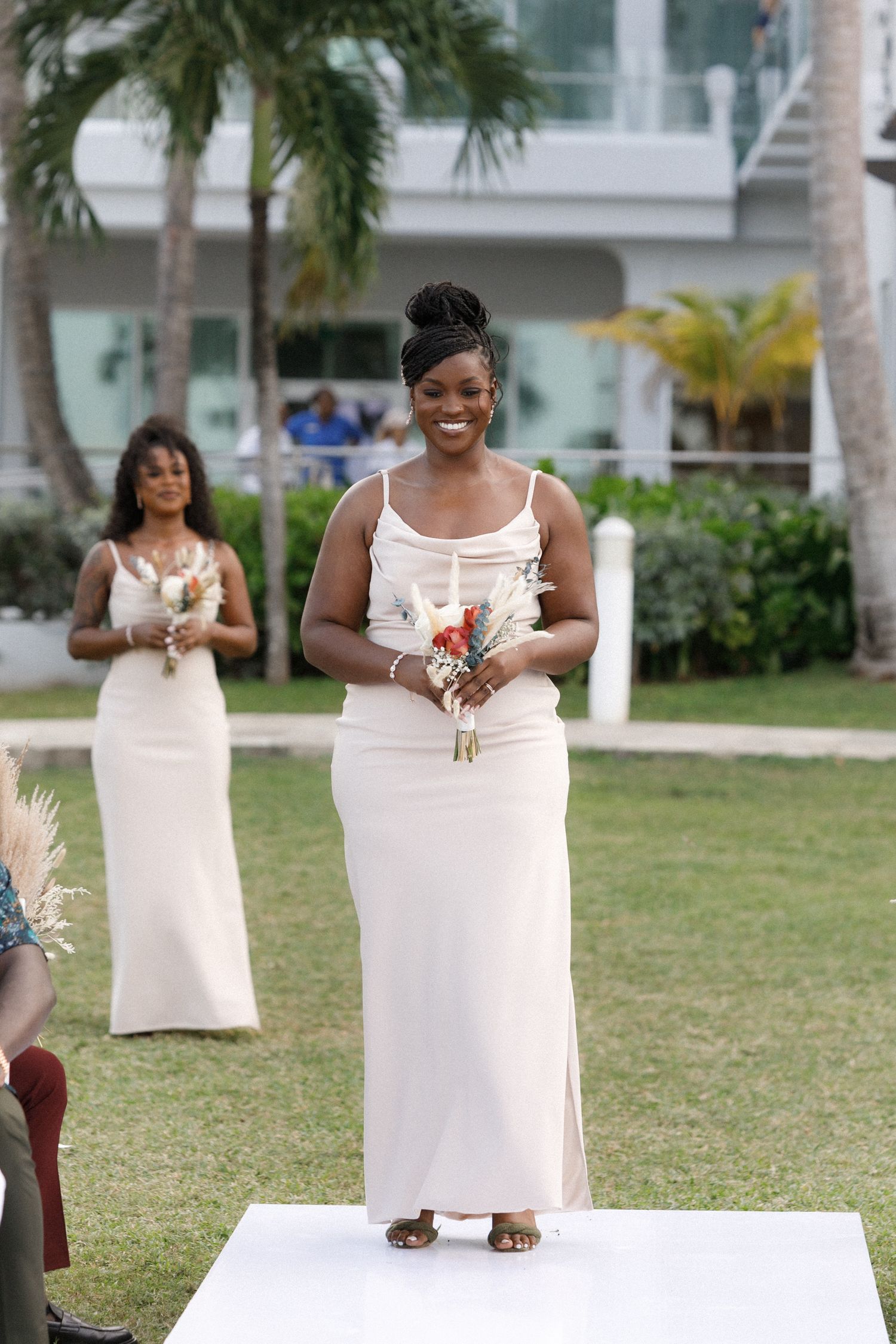 Members of a wedding party stand on a white runway during an outdoor ceremony with palm trees.