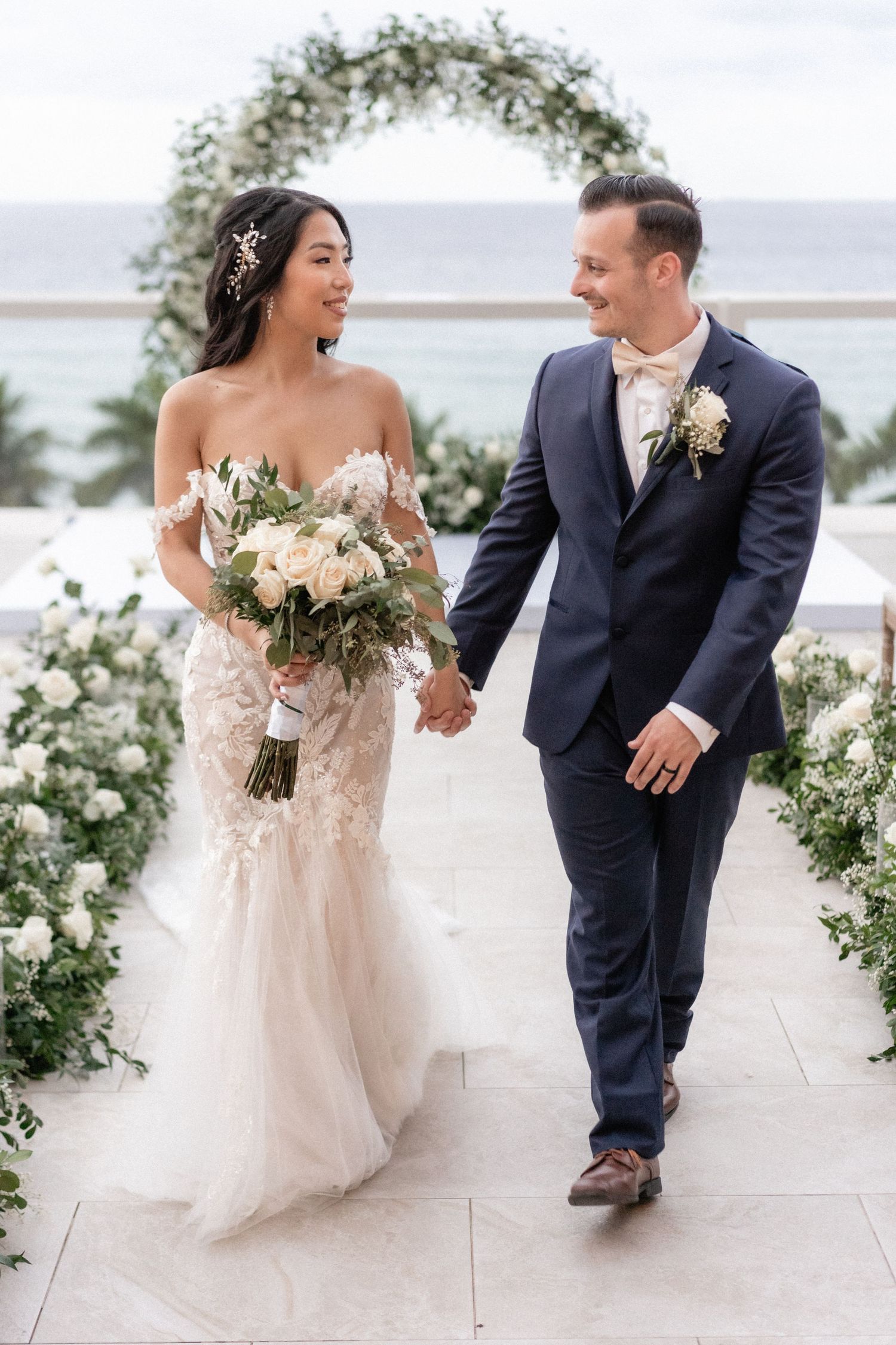 A wedding couple walking down an aisle decorated with white flowers and greenery under an outdoor arch by the ocean.
