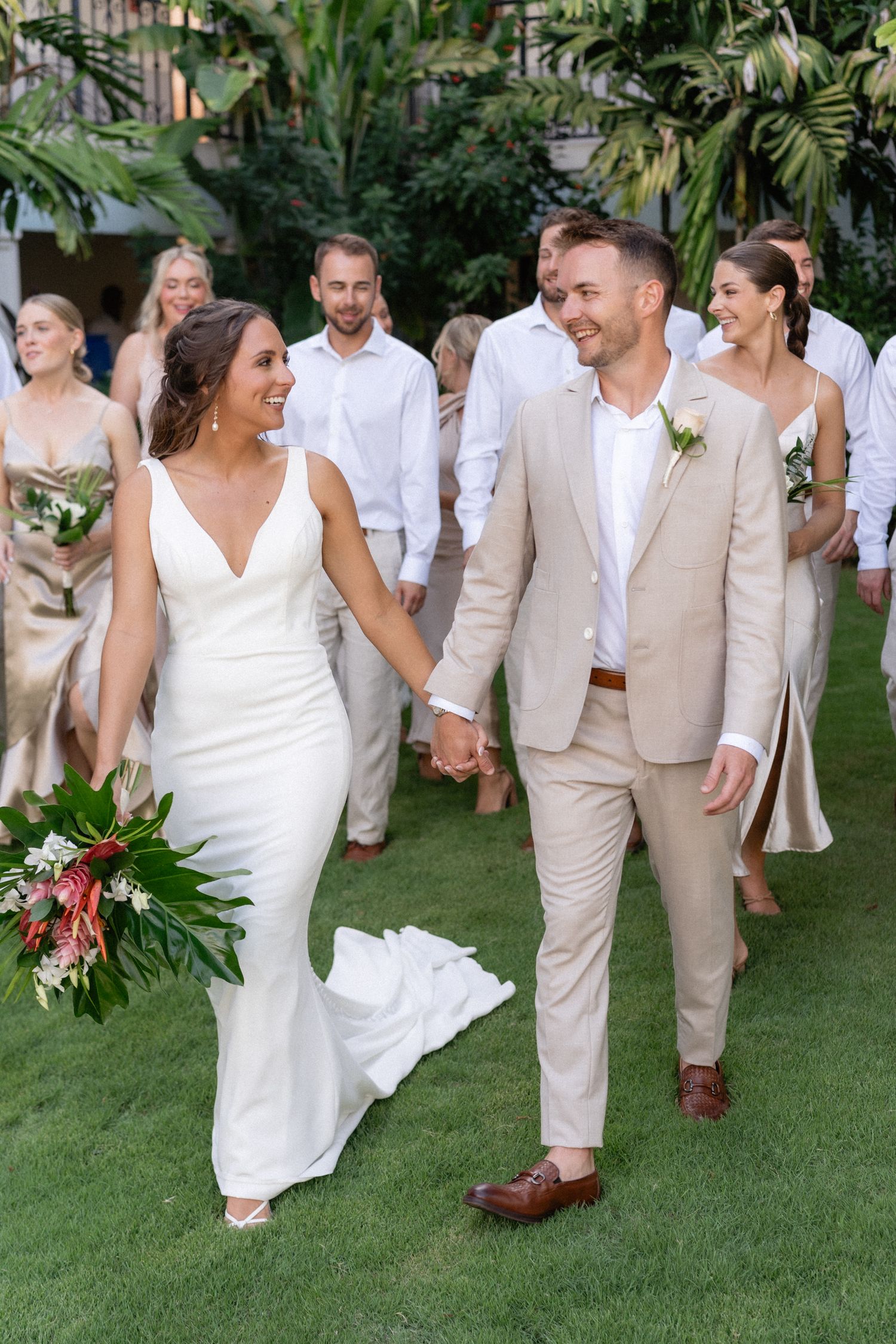 Wedding party walks on garden path with bride and groom in white dress and beige suit.