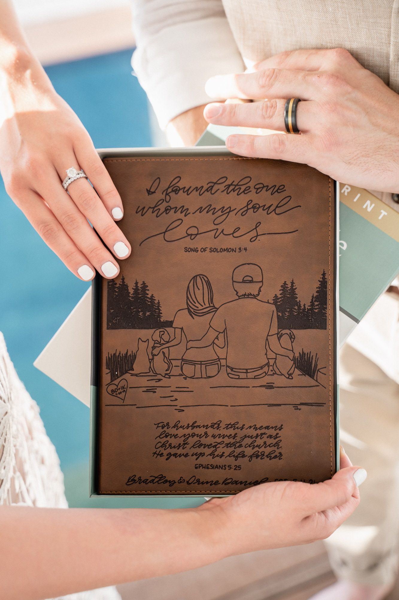 Hands holding a brown leather-bound book with an illustrated family scene and romantic quote engraved on the cover.