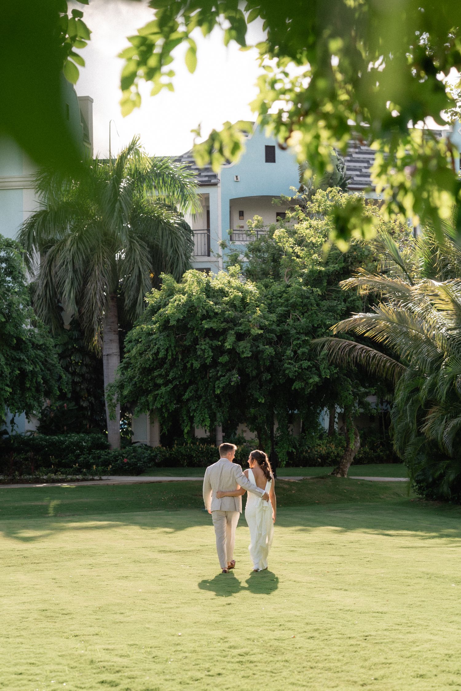 A couple stands together on a lush green lawn surrounded by tropical palm trees and resort buildings.