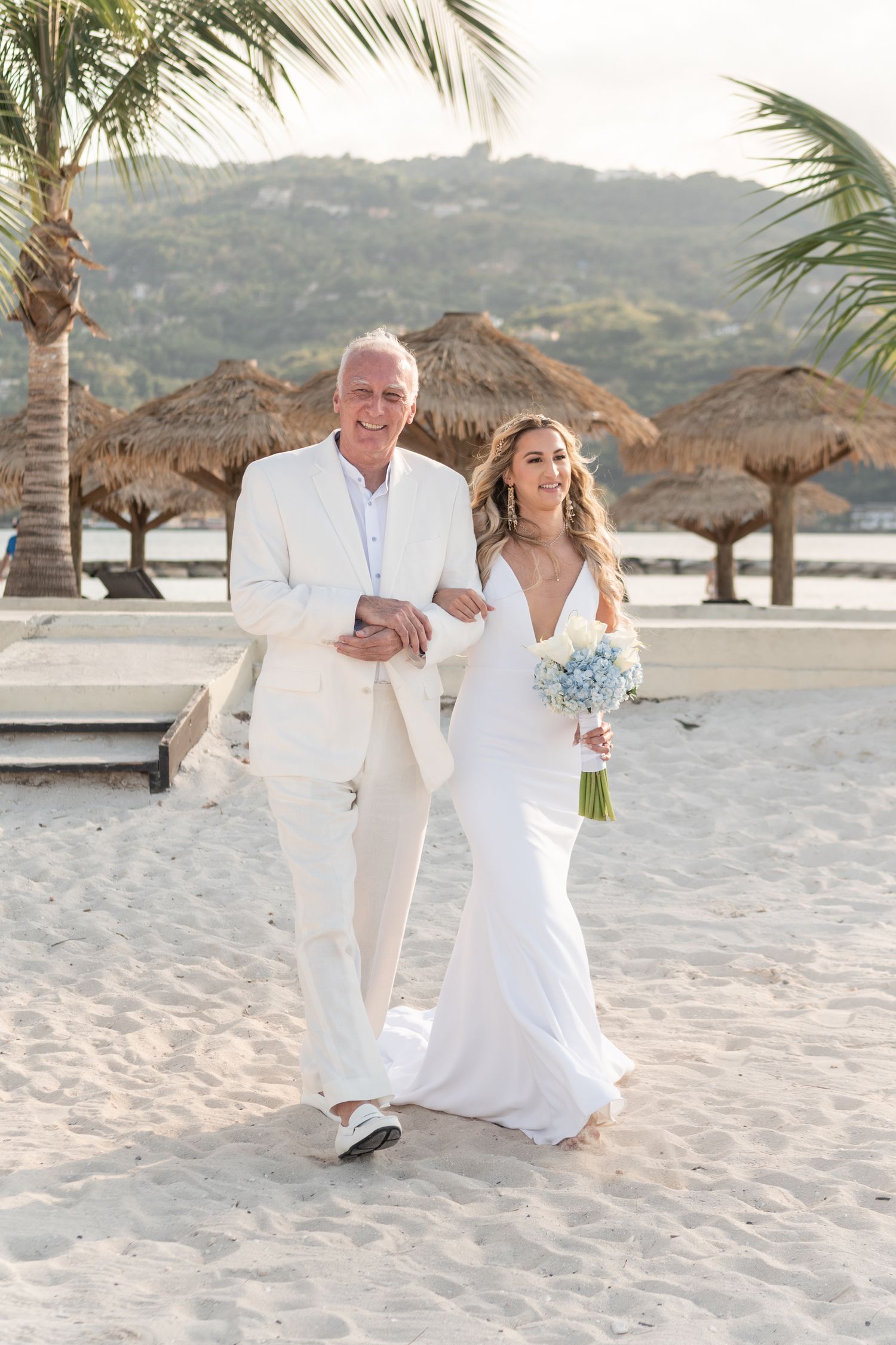 Couple walking on sandy beach with thatched umbrellas and palm trees at destination wedding.