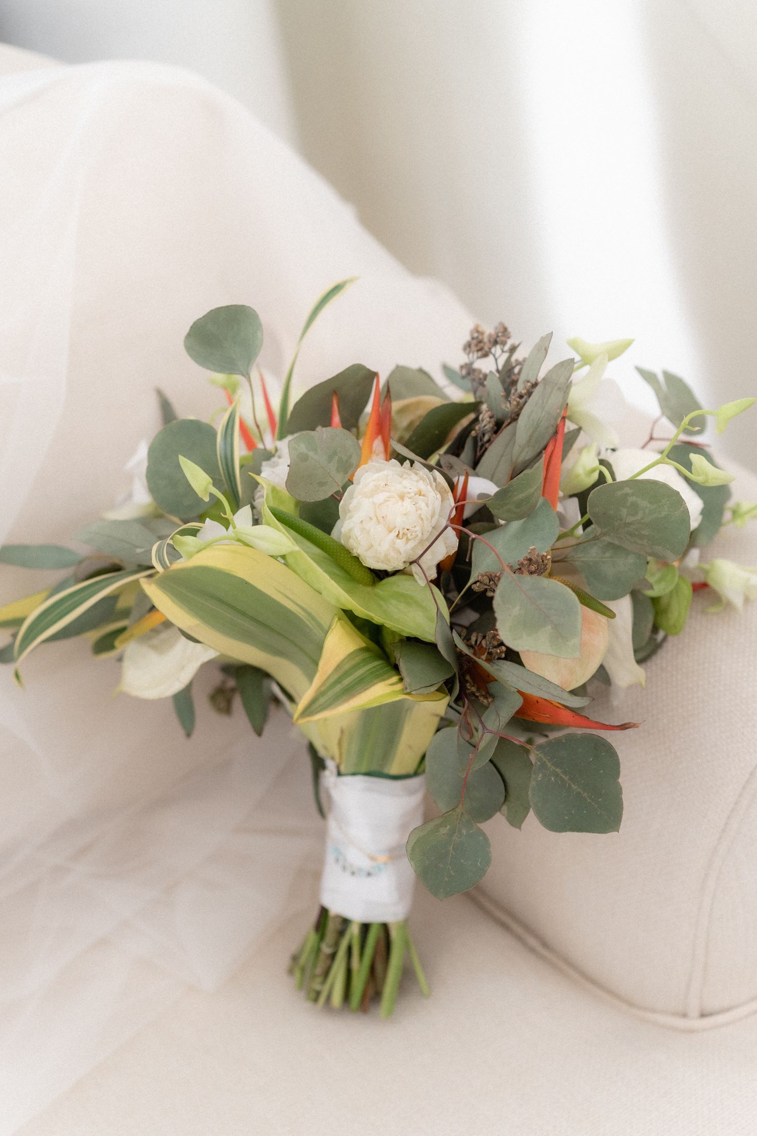 An elegant wedding bouquet with white flowers, eucalyptus leaves and orange tropical accents on a white chair.