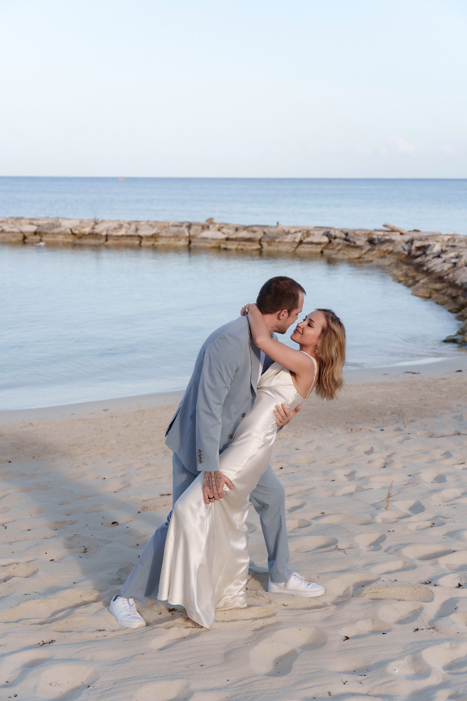 A romantic beach wedding photo of a couple in white dress and grey suit sharing a passionate kiss by the ocean.