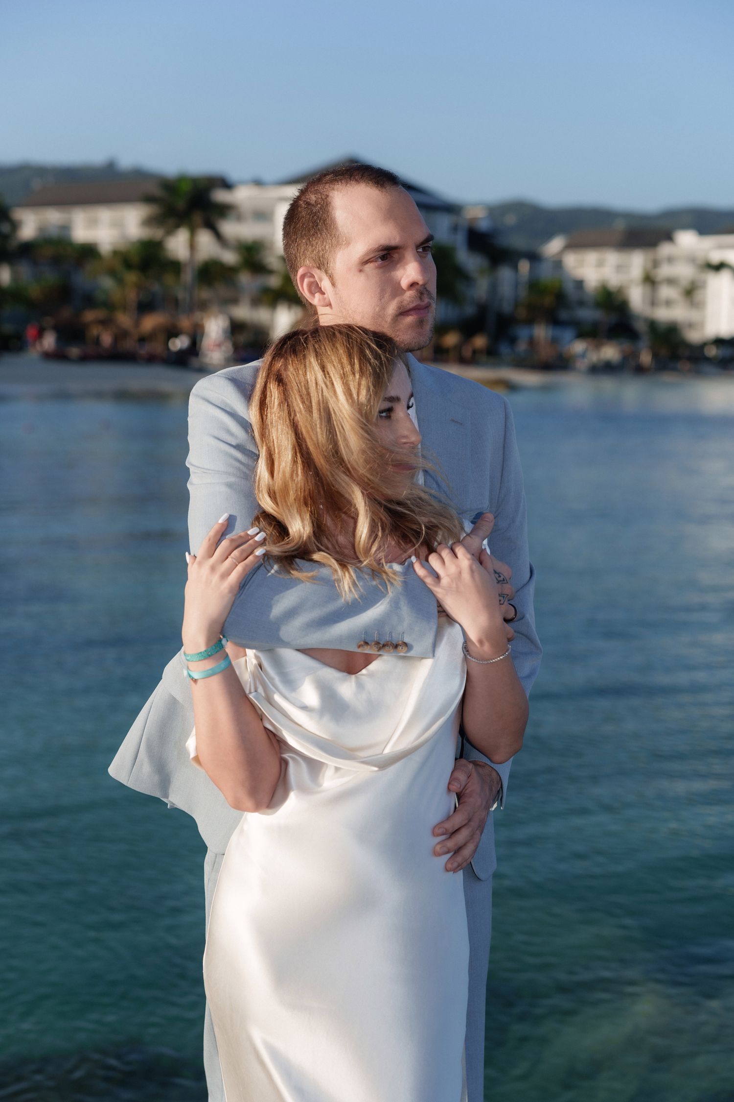 Romantic embrace on oceanfront with resort buildings visible in background.