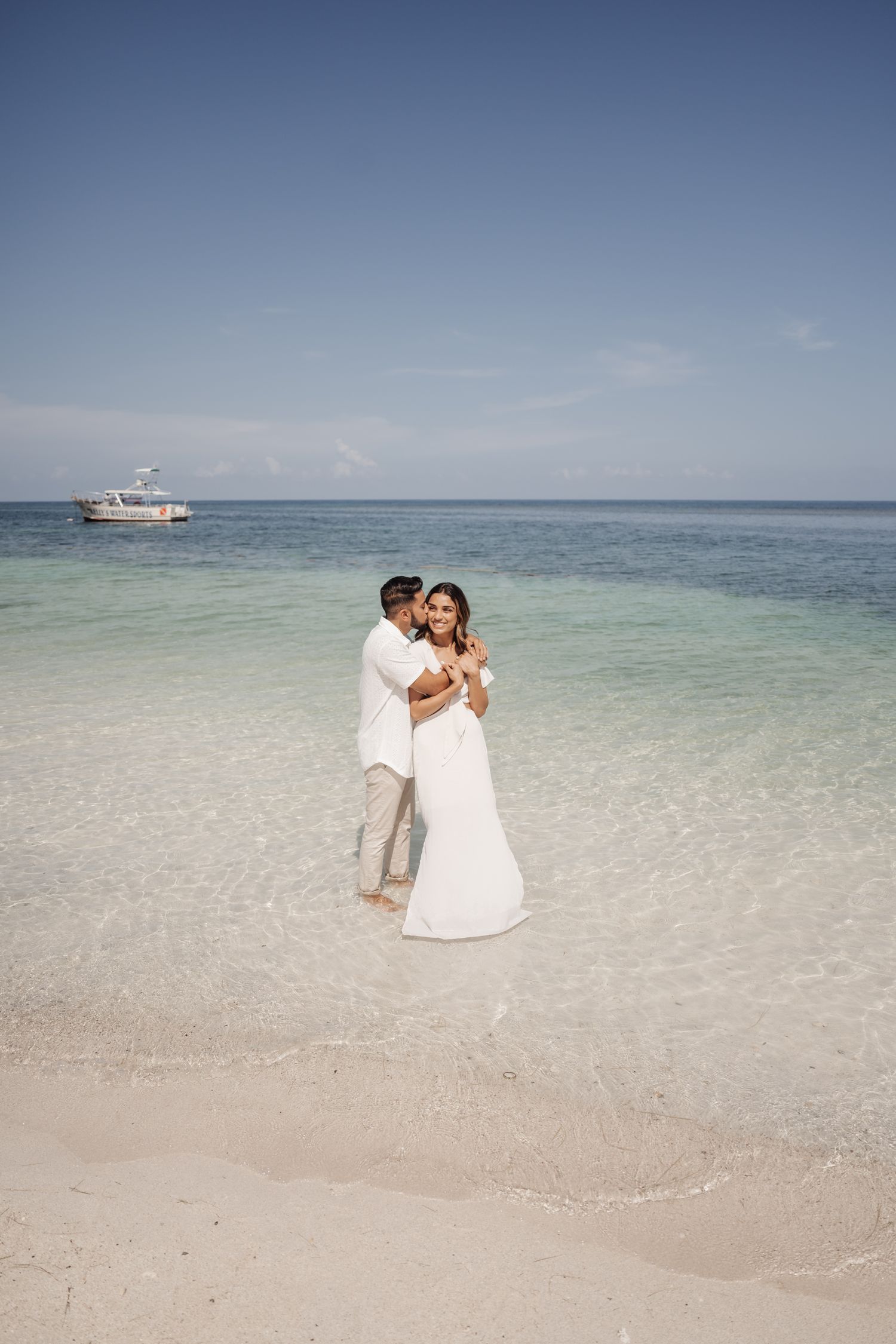 A romantic couple embraces on a pristine beach with crystal clear waters and a boat in the distance.