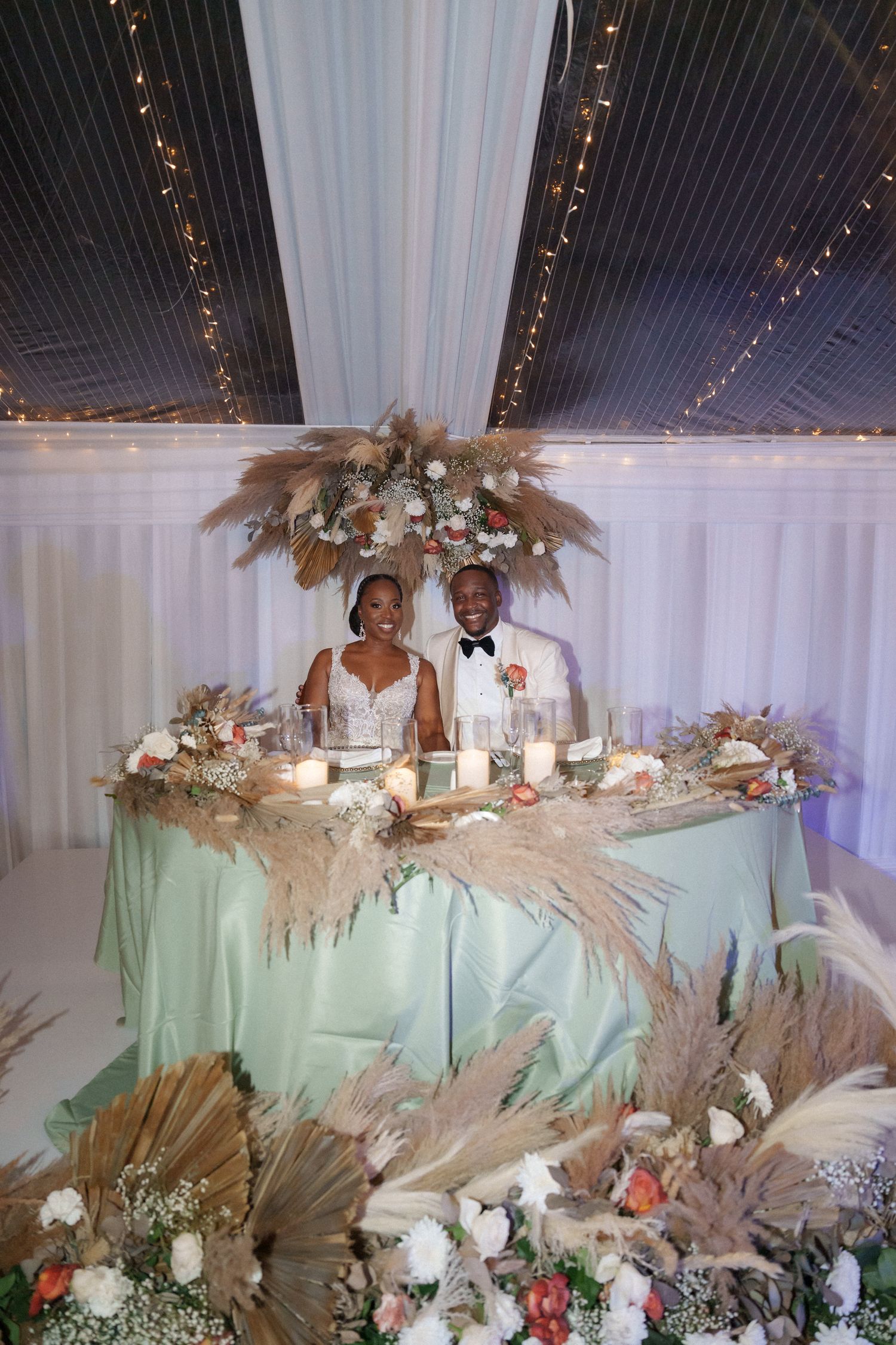 Wedding sweetheart table decorated with pampas grass and floral arrangements under a white draped ceiling.