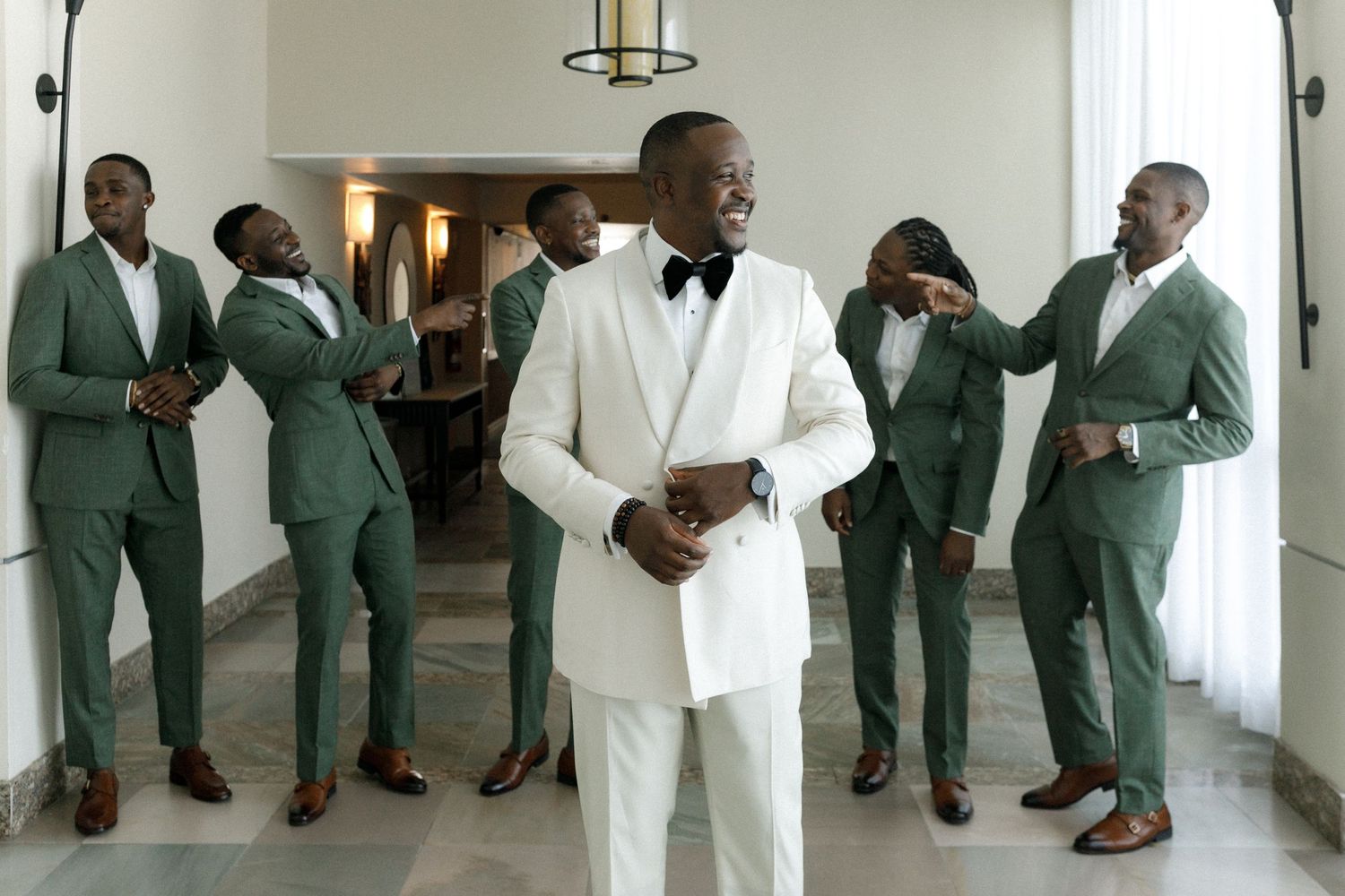 Groomsmen in sage green suits gather around groom in white tuxedo during wedding preparations.