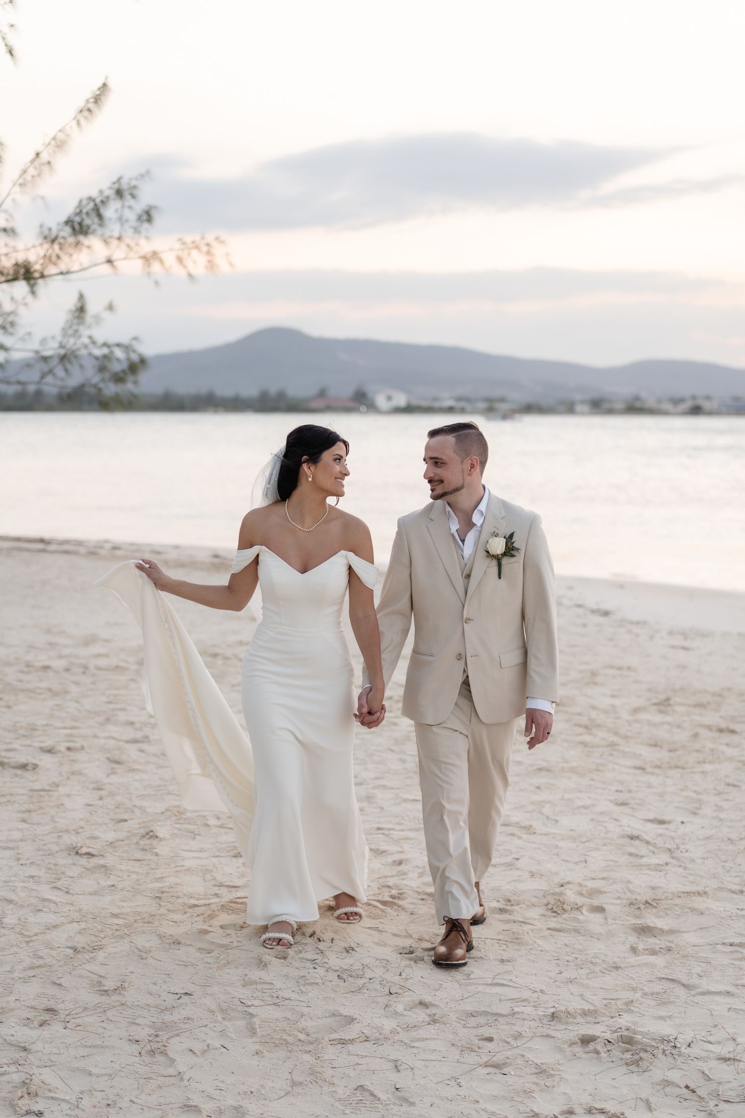 A couple walks hand in hand on a beach at sunset in elegant wedding attire.