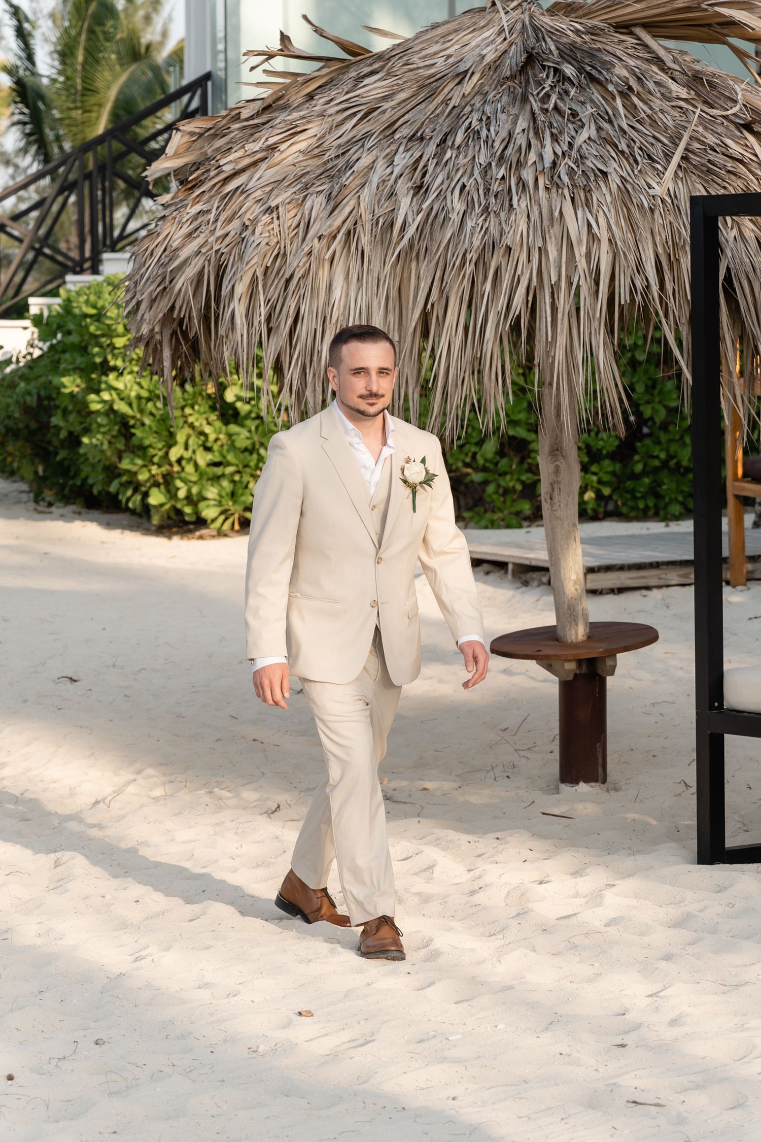 Wedding attire in beige suit walking along tropical beach path under palm fronds.
