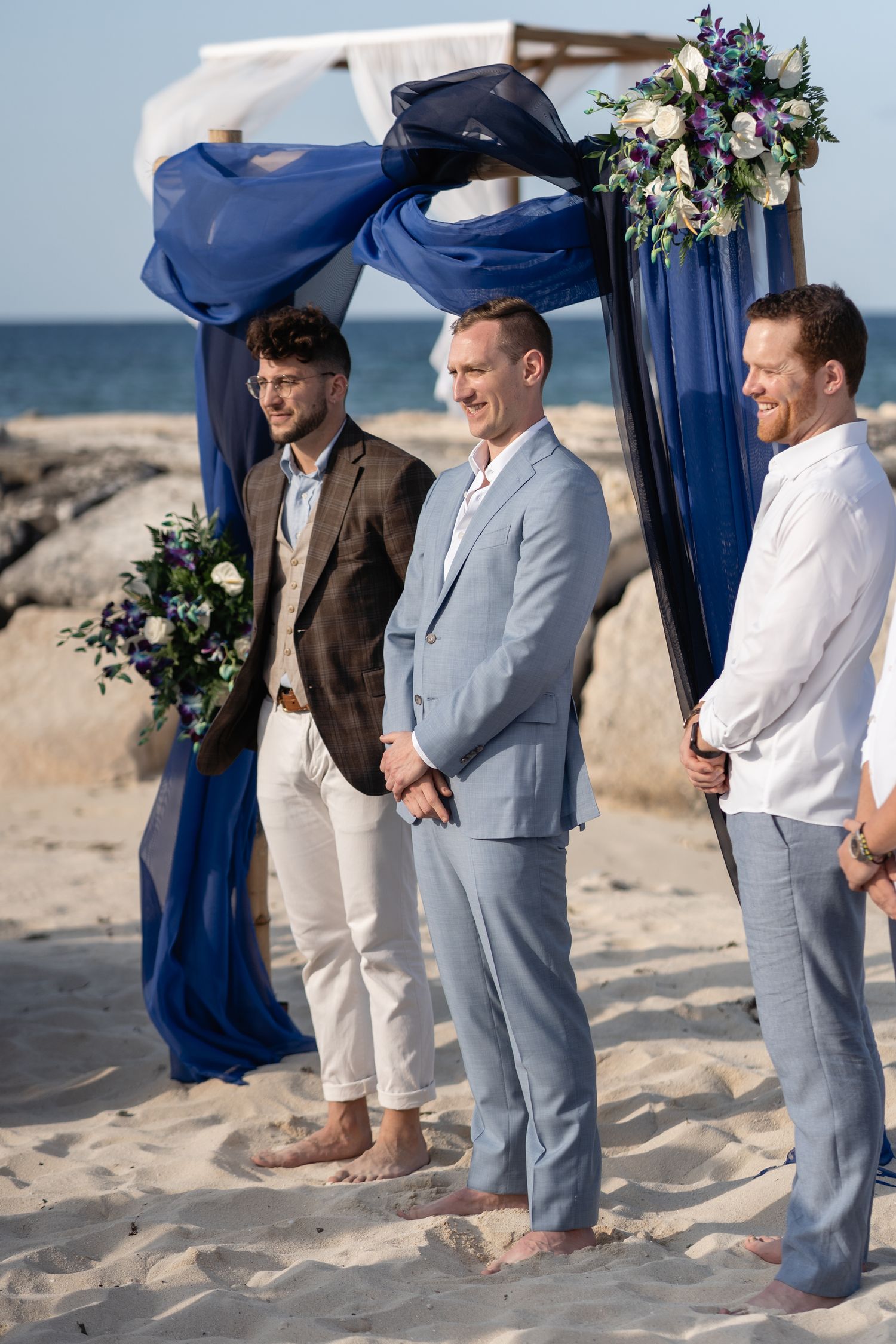 Three groomsmen standing barefoot on beach under navy blue arch with floral arrangements.