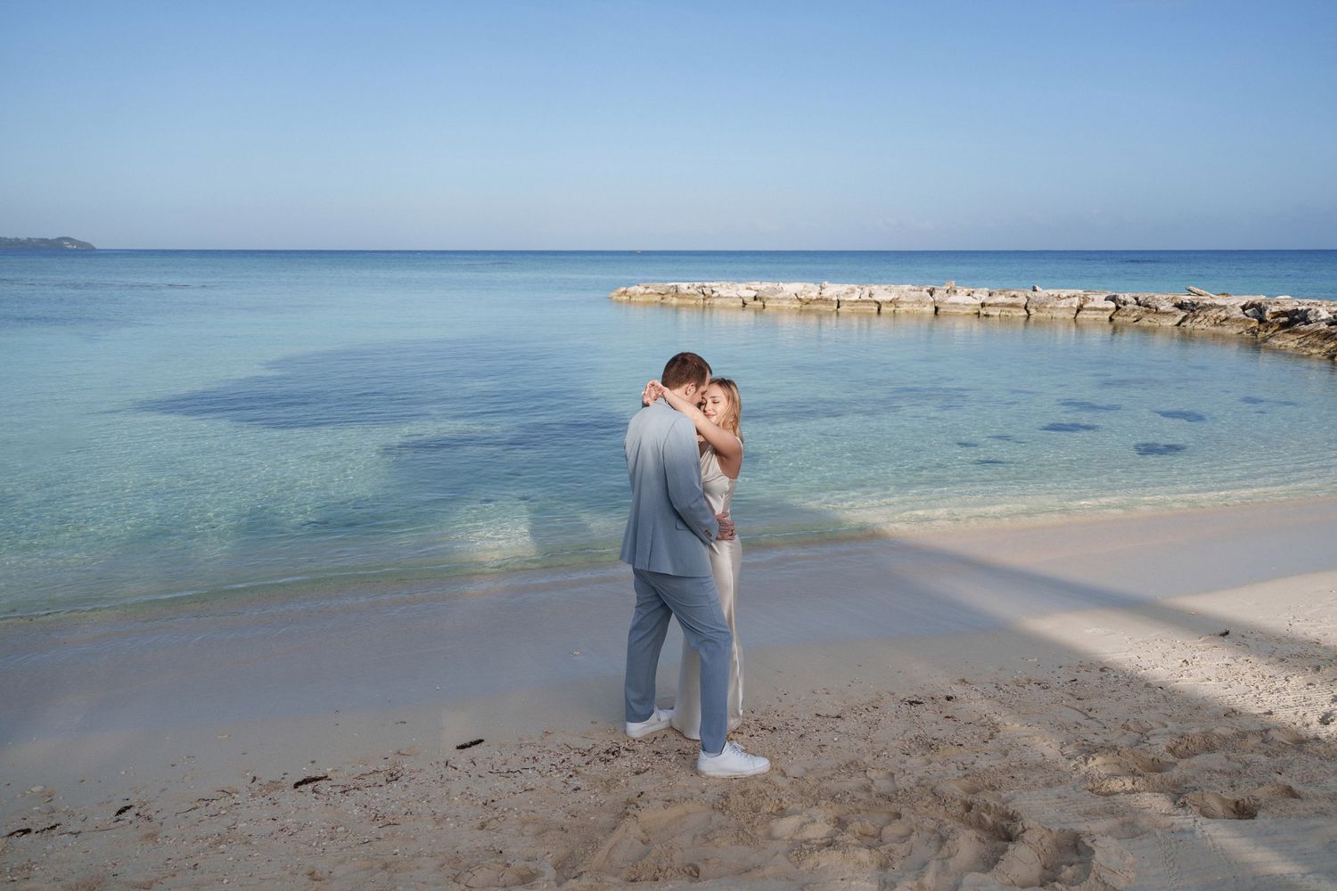 A romantic moment captured between two people embracing on a pristine beach with turquoise waters and a stone jetty.