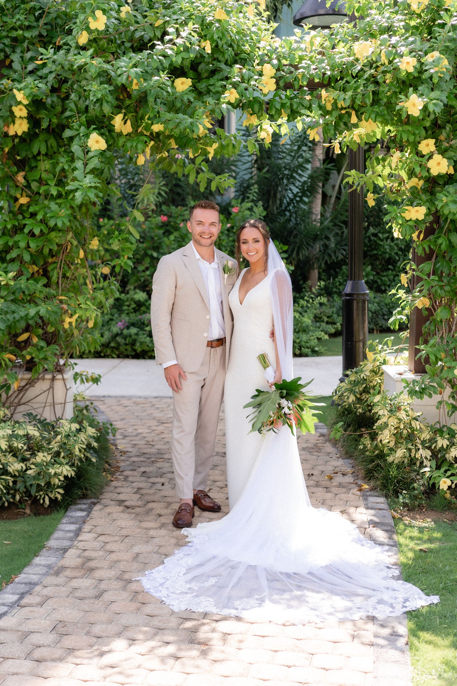 Wedding couple standing on a stone pathway surrounded by yellow flowering vines and tropical gardens.