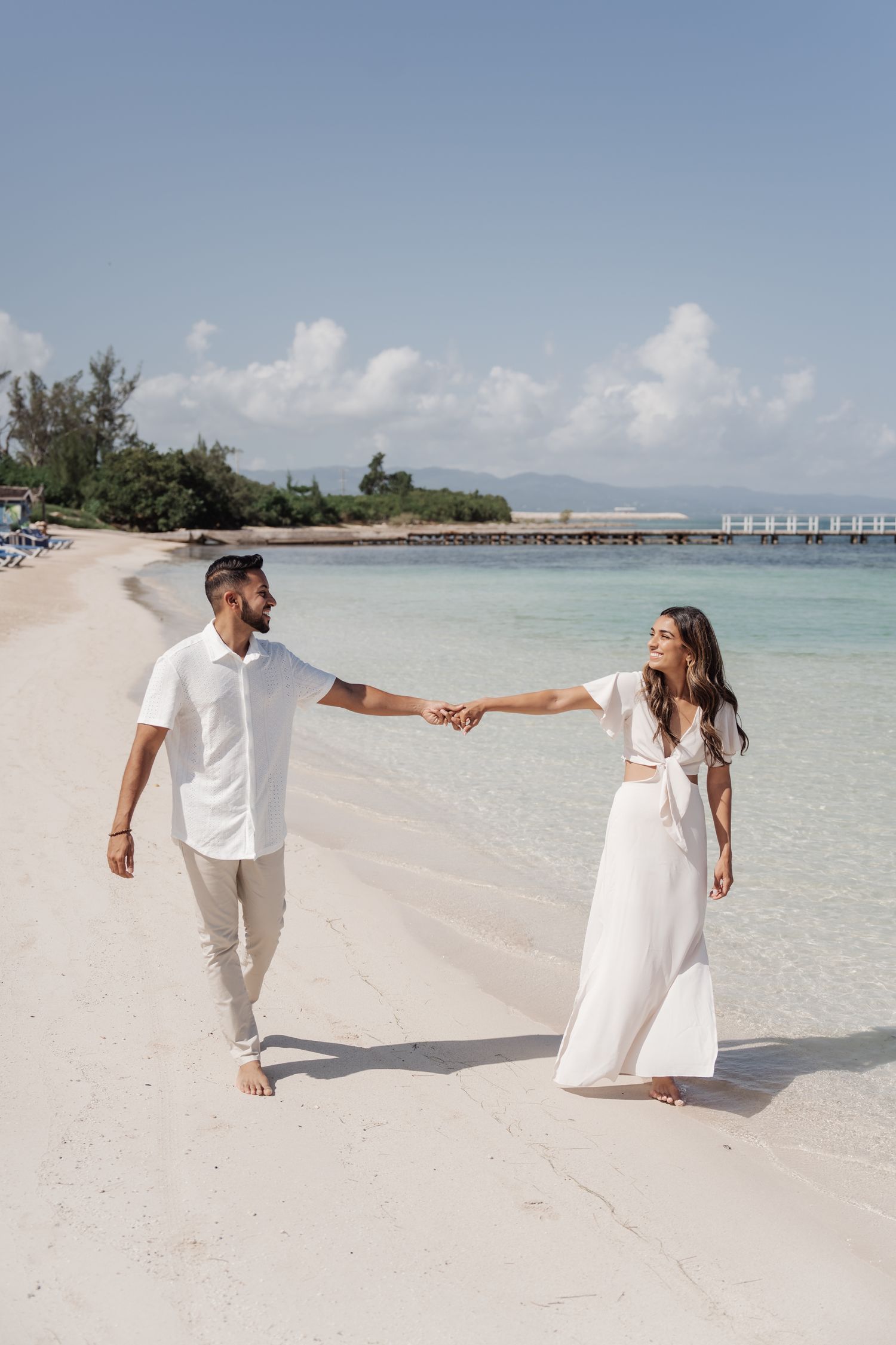 Couple in white outfits holding hands while walking on tropical beach.