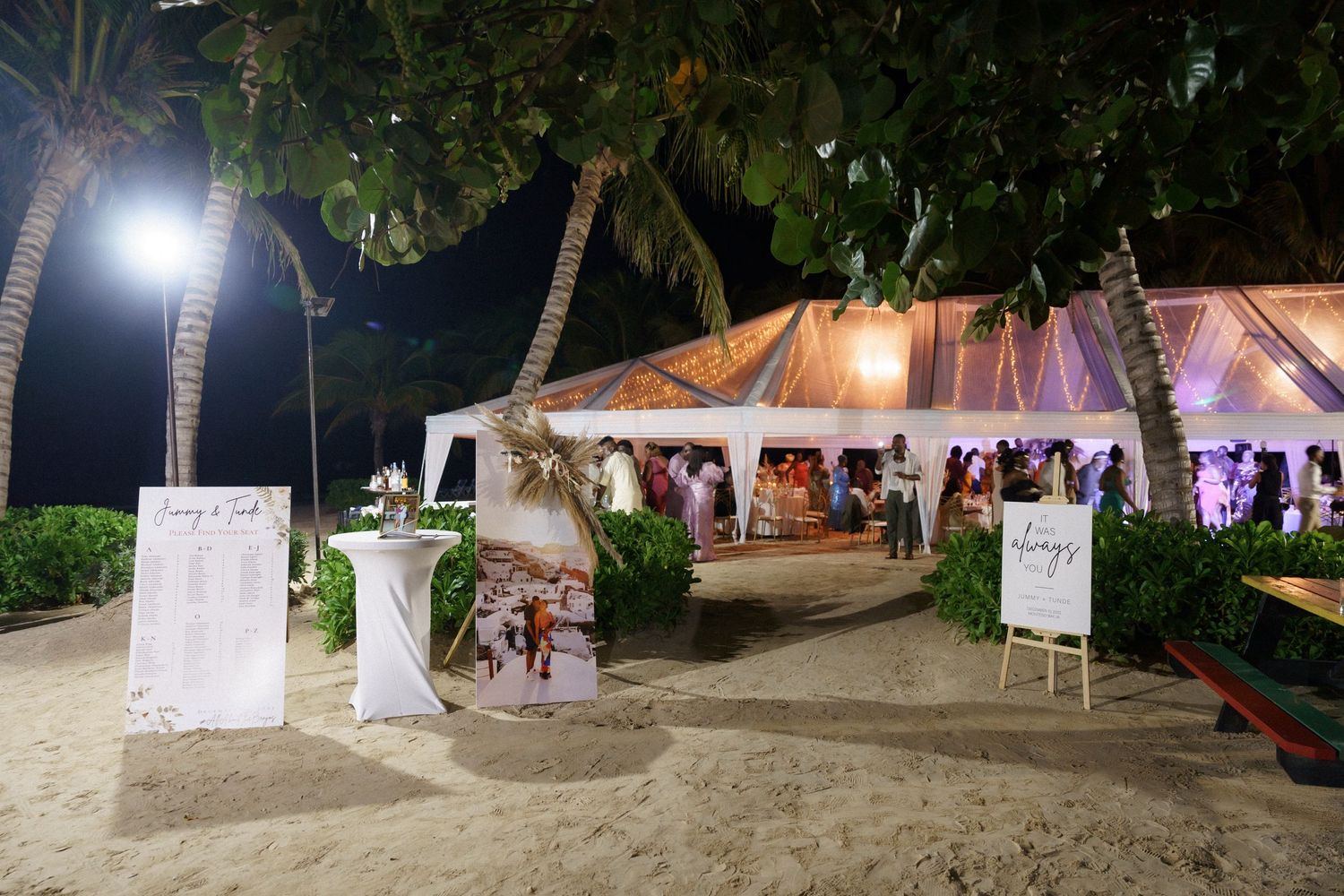 Outdoor beach wedding reception tent with illuminated palm trees and welcome signs.