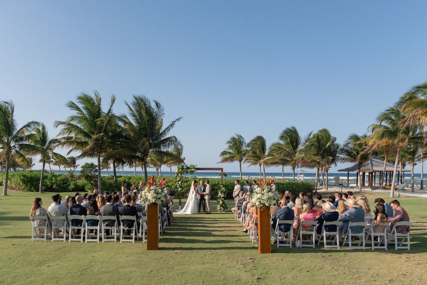 Outdoor wedding ceremony setup with white chairs arranged on a lawn lined with palm trees by the ocean.