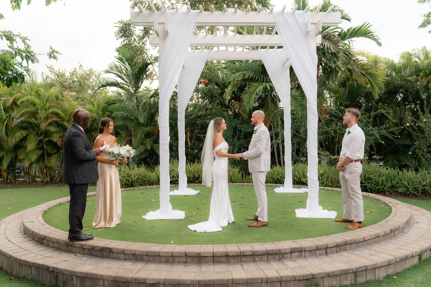 Intimate wedding ceremony under a white draped gazebo surrounded by tropical gardens.