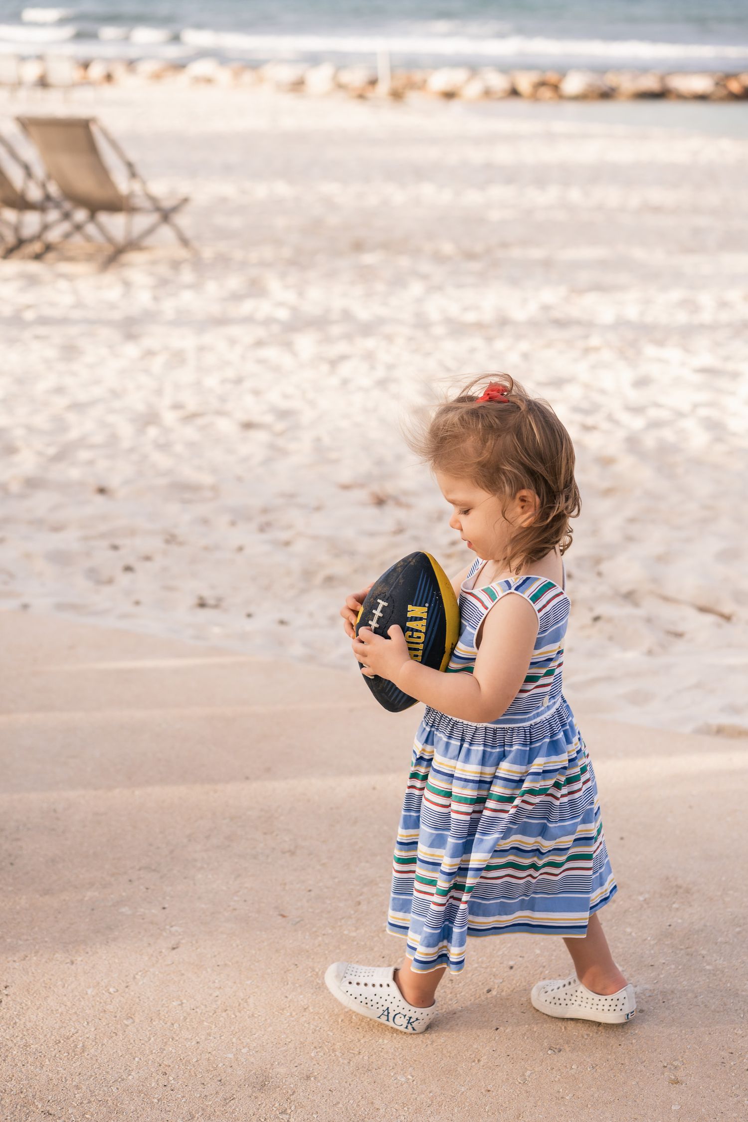 Small child in striped dress walking on beach while holding football.