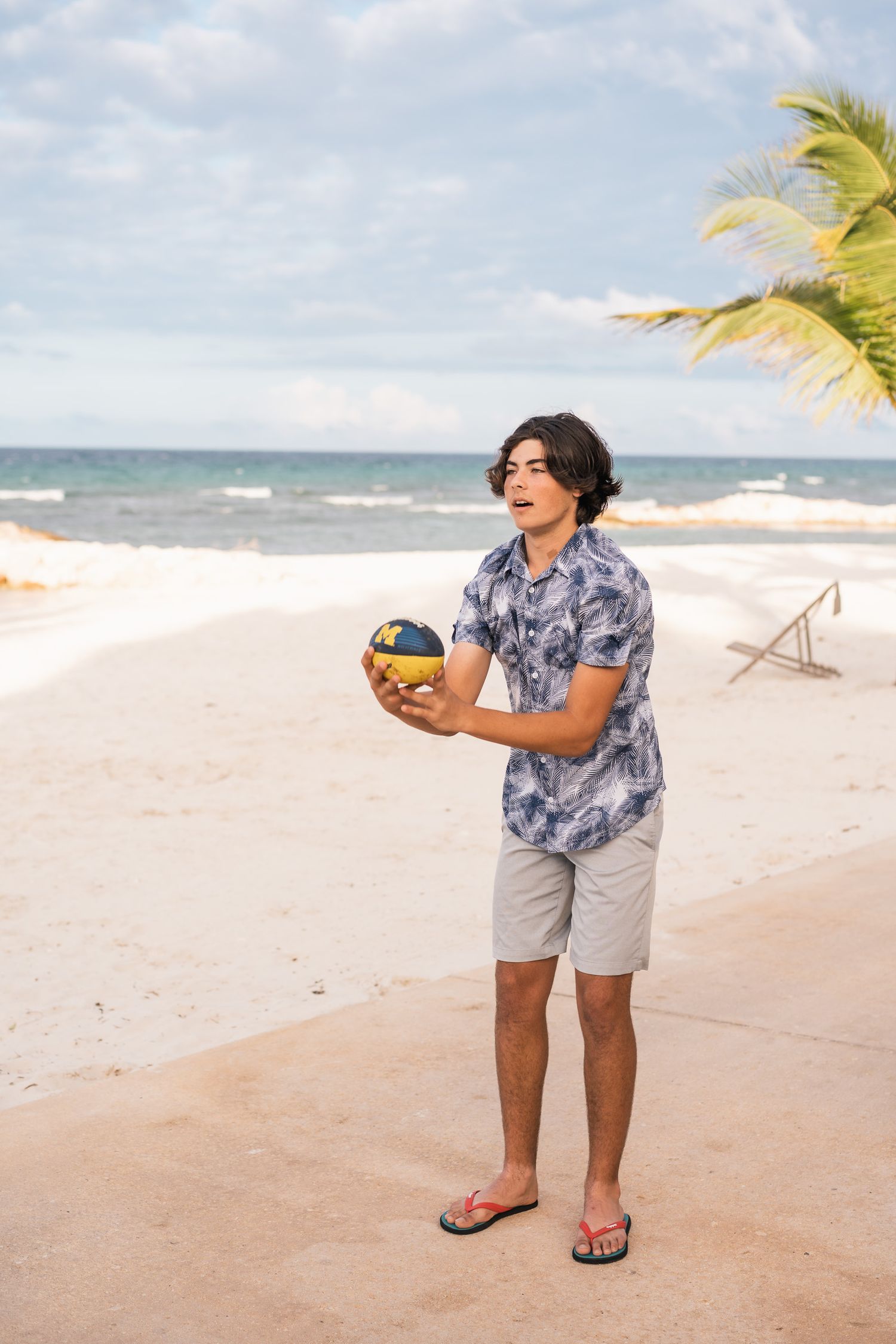 Person in floral shirt holding volleyball on sunny tropical beach with palm trees.