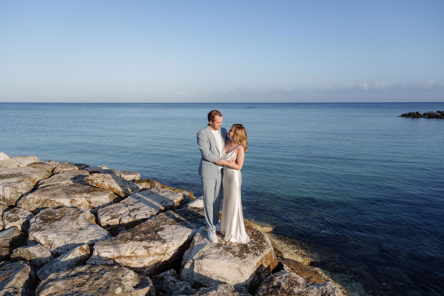 Couple in wedding attire standing on coastal rocks overlooking the ocean.