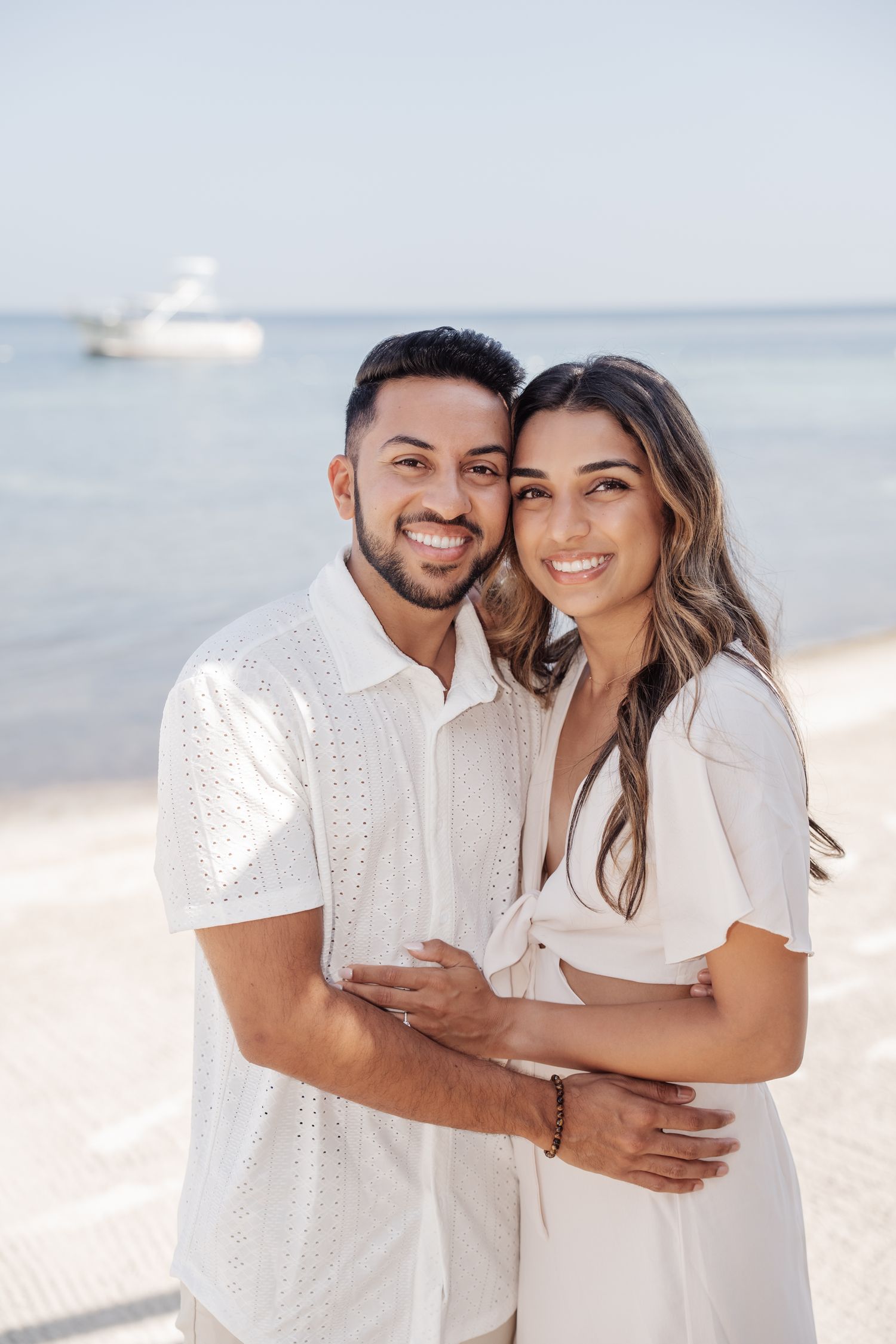 A happy couple poses on a sunny beach with a boat visible in the distance.