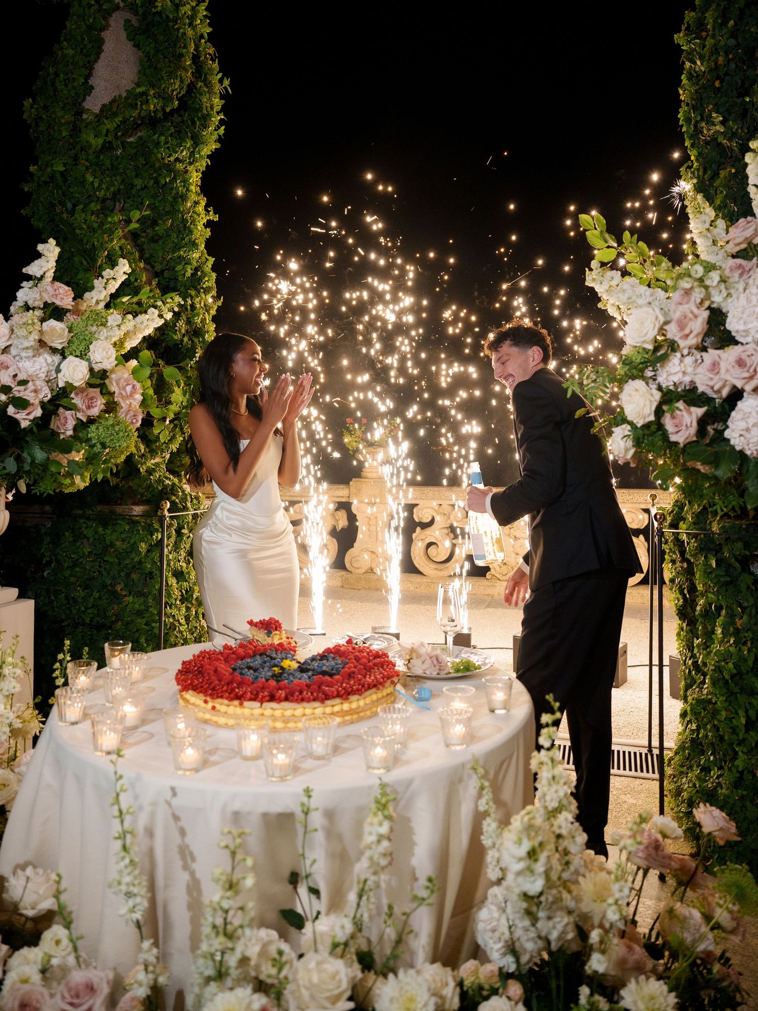 Wedding cake display surrounded by sparklers and floral decorations creating a magical evening ambiance.