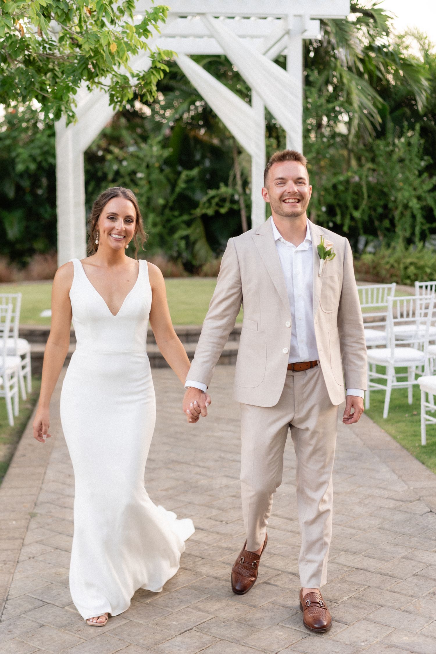 Newlyweds walk together under white pergola with garden ceremony setup.