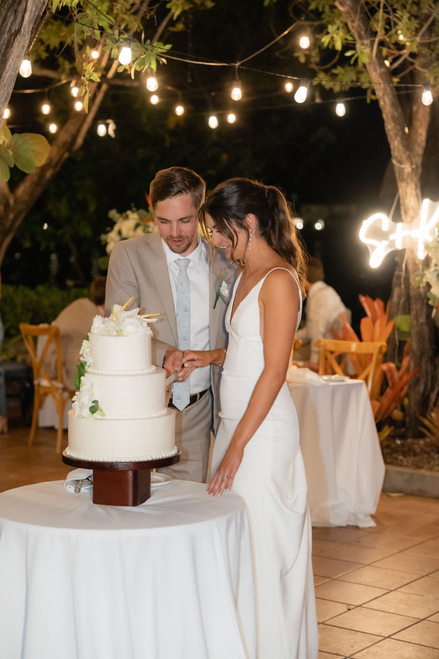 Couple cuts elegant white wedding cake under string lights at outdoor evening reception.