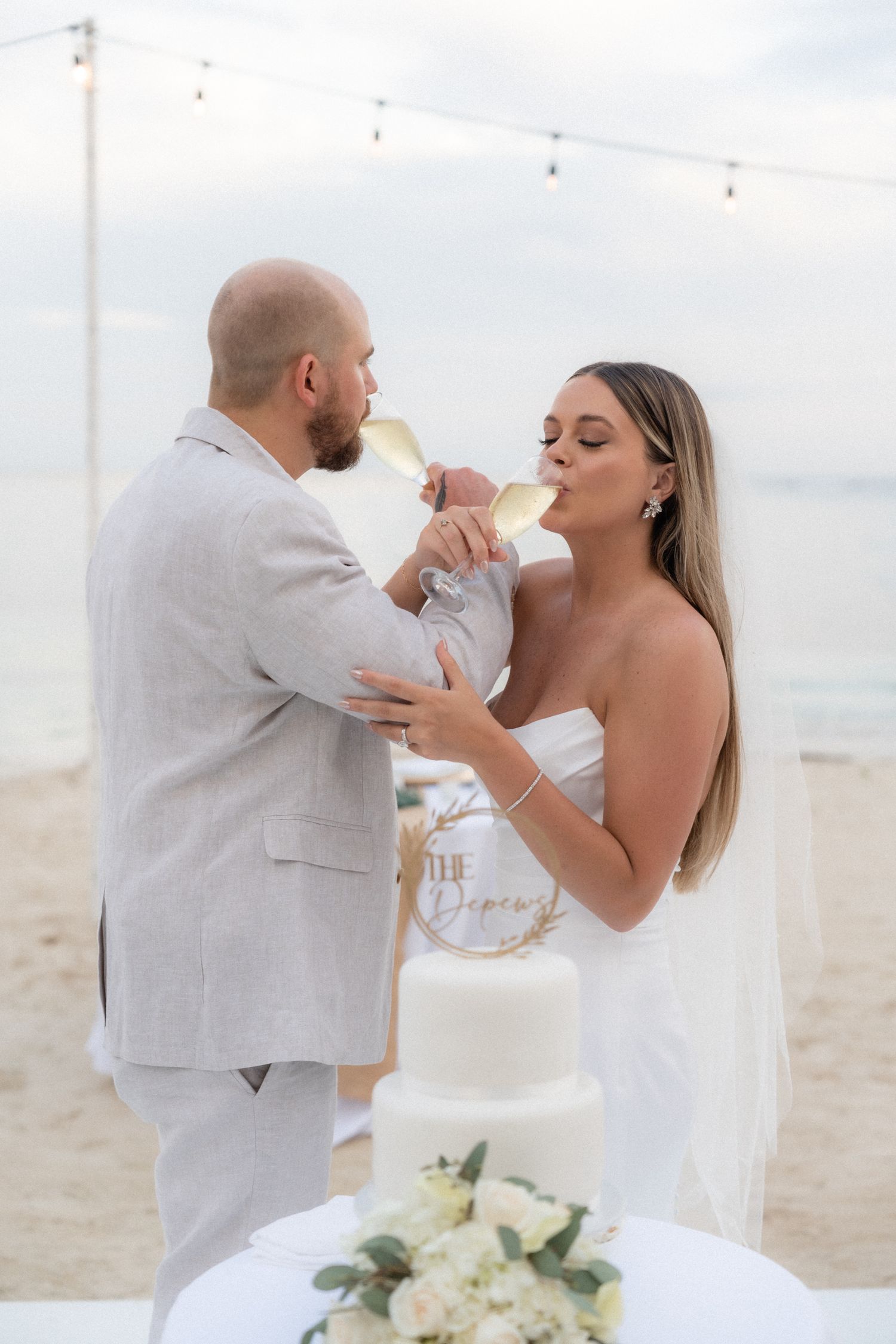 Newlyweds share champagne toast on beach under string lights at sunset.