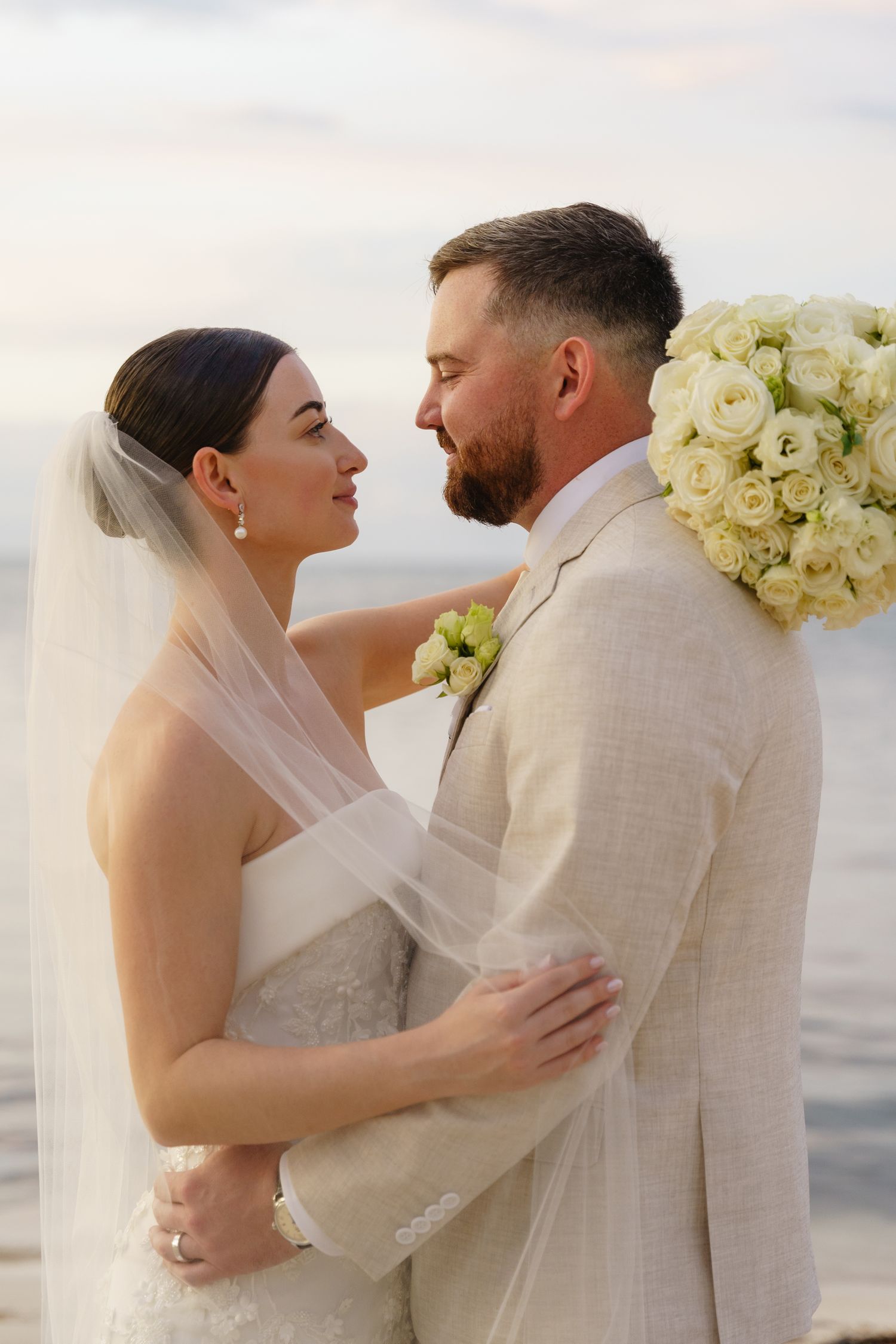 A tender moment between newlyweds on the beach at sunset with a large white rose bouquet visible.