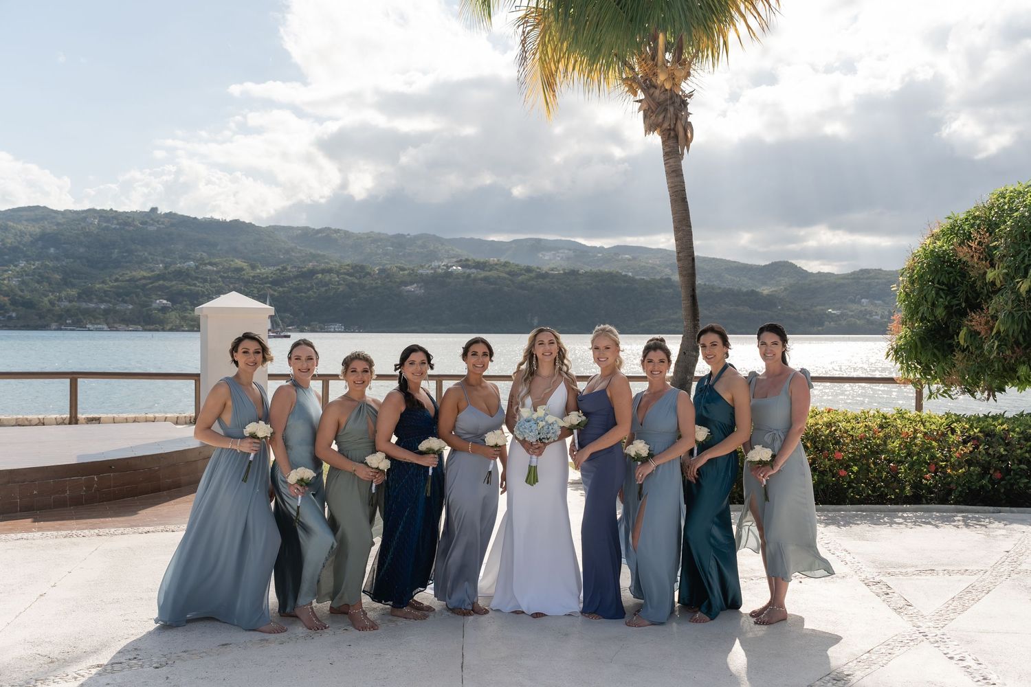 Wedding party in coordinating blue and gray dresses poses by the water under palm trees.