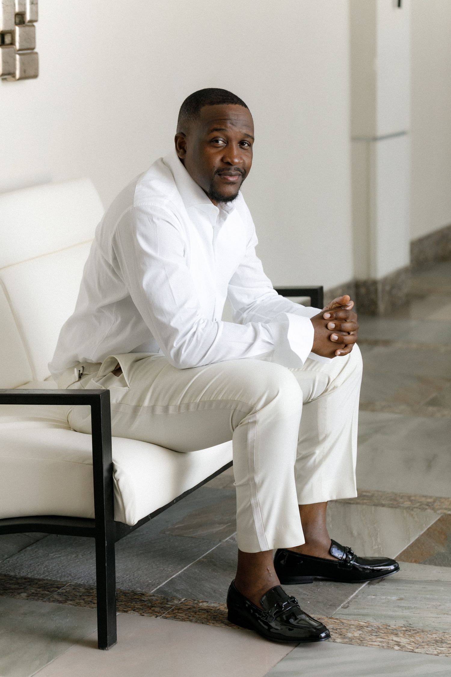 Groom in all-white attire sitting on modern chair in bright interior setting.