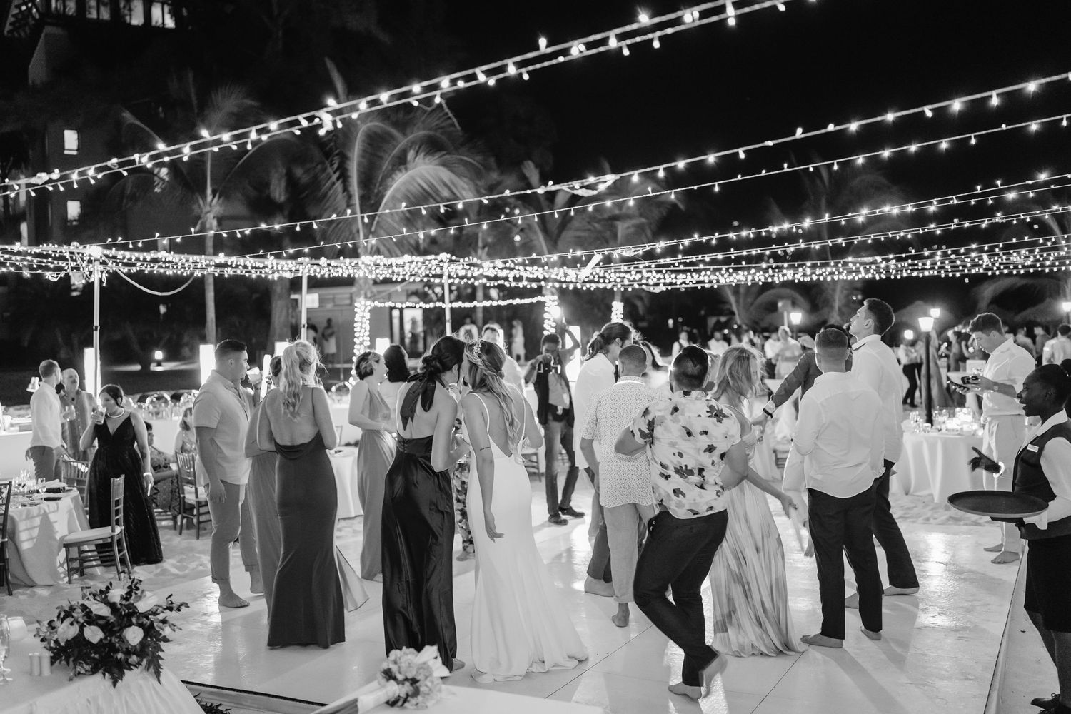 Black and white photo of wedding guests dancing under string lights at an evening reception.