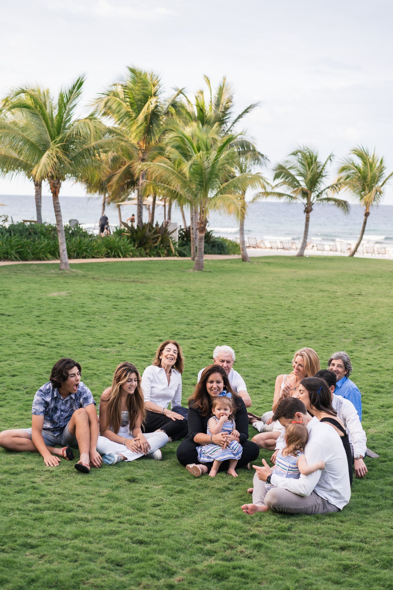 Family gathering on oceanfront lawn with palm trees at tropical resort.
