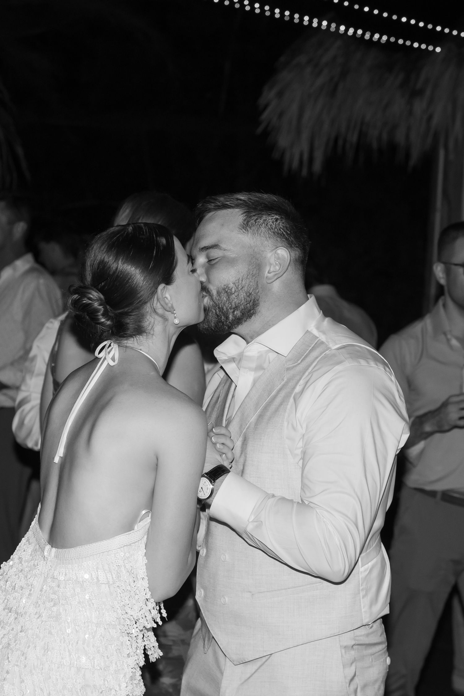 A romantic black and white photo of a couple sharing a kiss at what appears to be their wedding celebration.