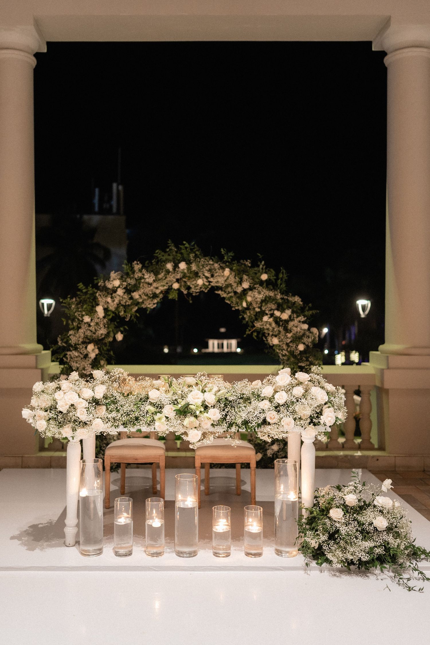An elegant floral arch with white roses decorates a wedding sweetheart table at night.