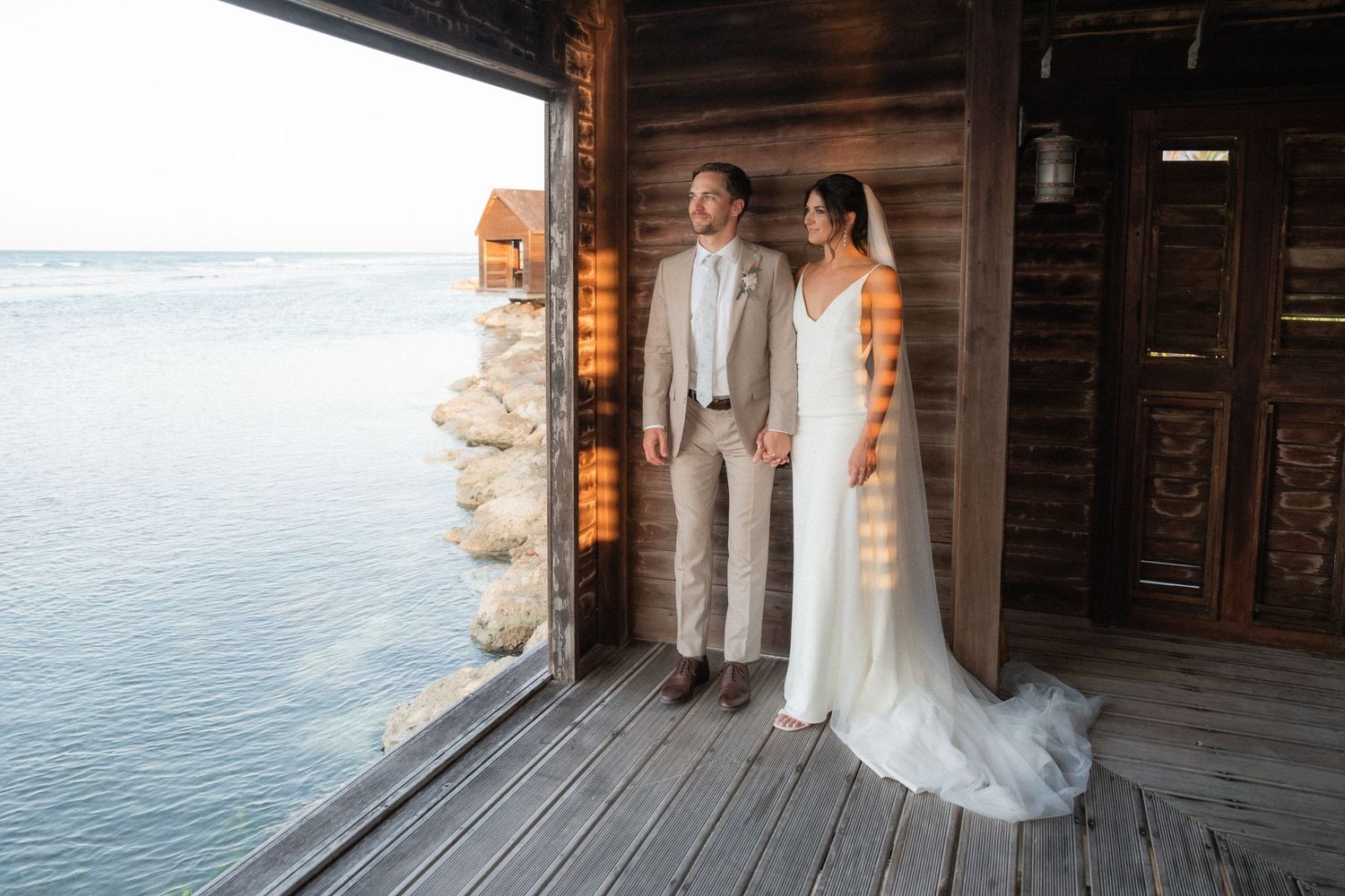 Wedding couple standing on a wooden deck overlooking the ocean at sunset.