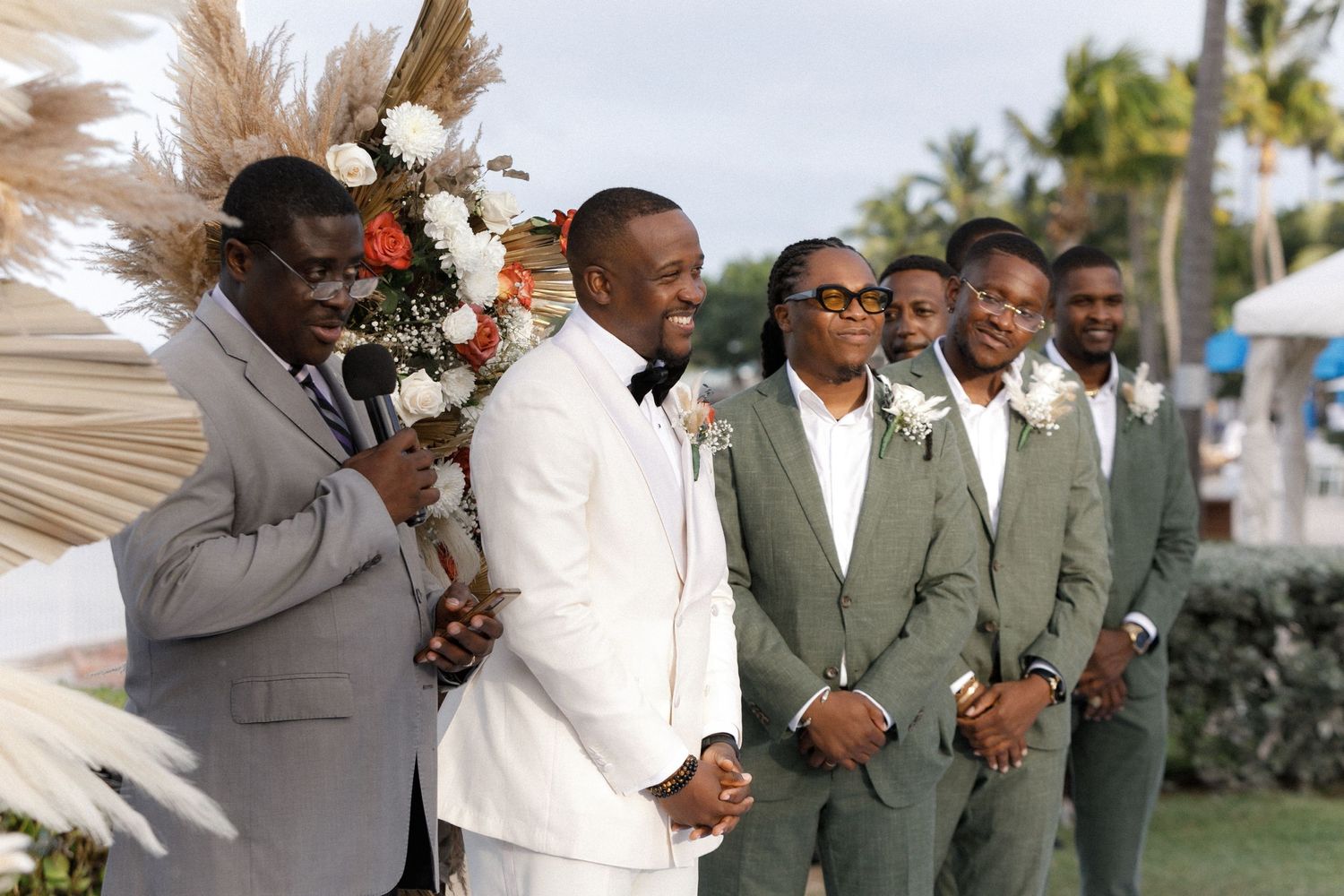 Groomsmen in green suits stand with the groom in white during an outdoor tropical wedding ceremony.