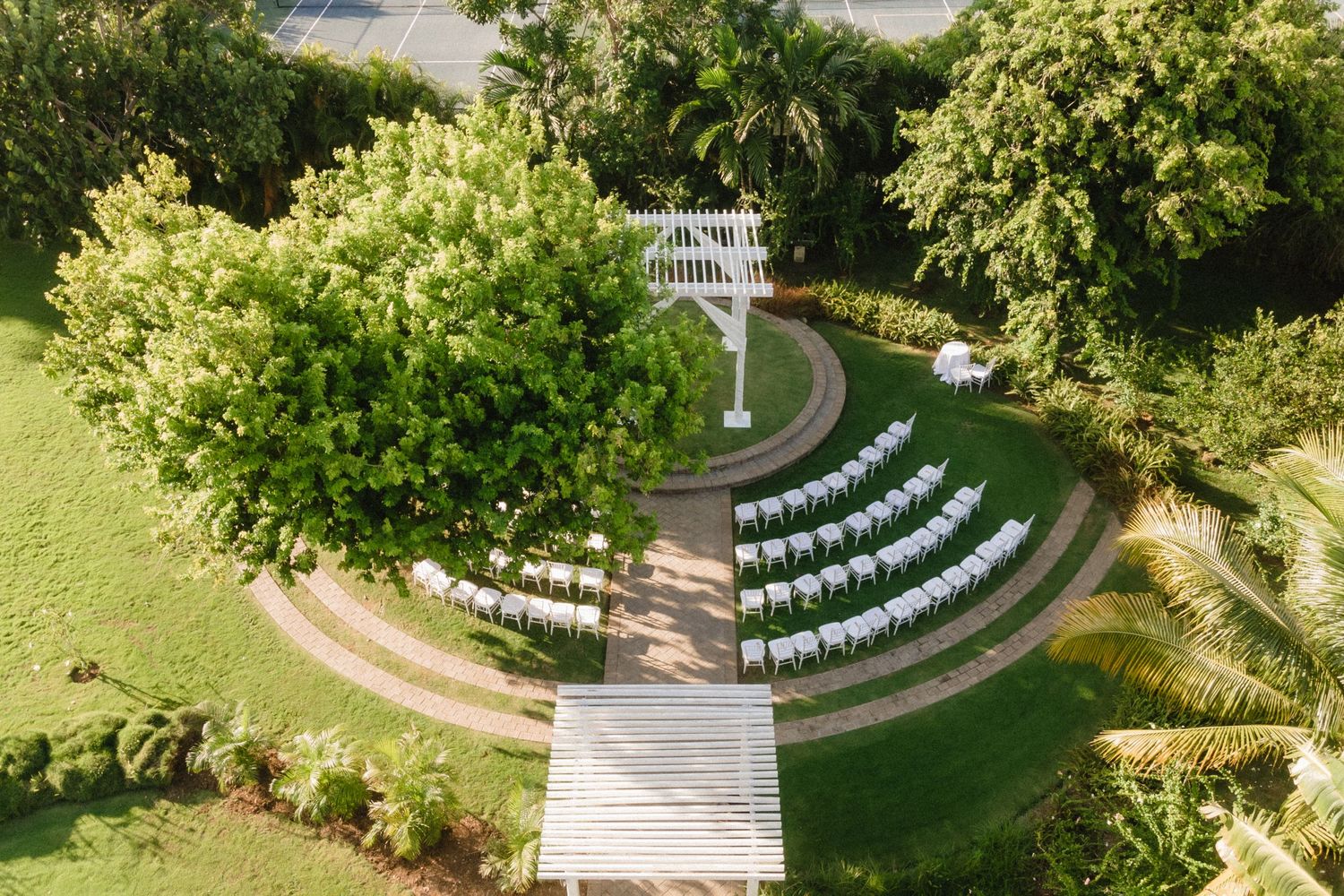 Aerial view of curved rows of white chairs surrounding garden wedding pergola in tropical setting.