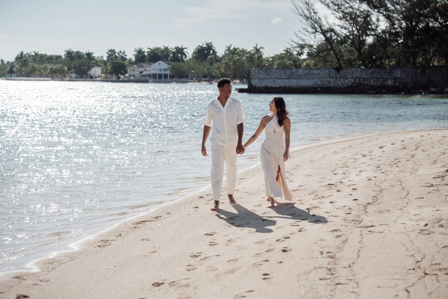 A couple walks hand in hand along a sunlit beach with sparkling water in the background.